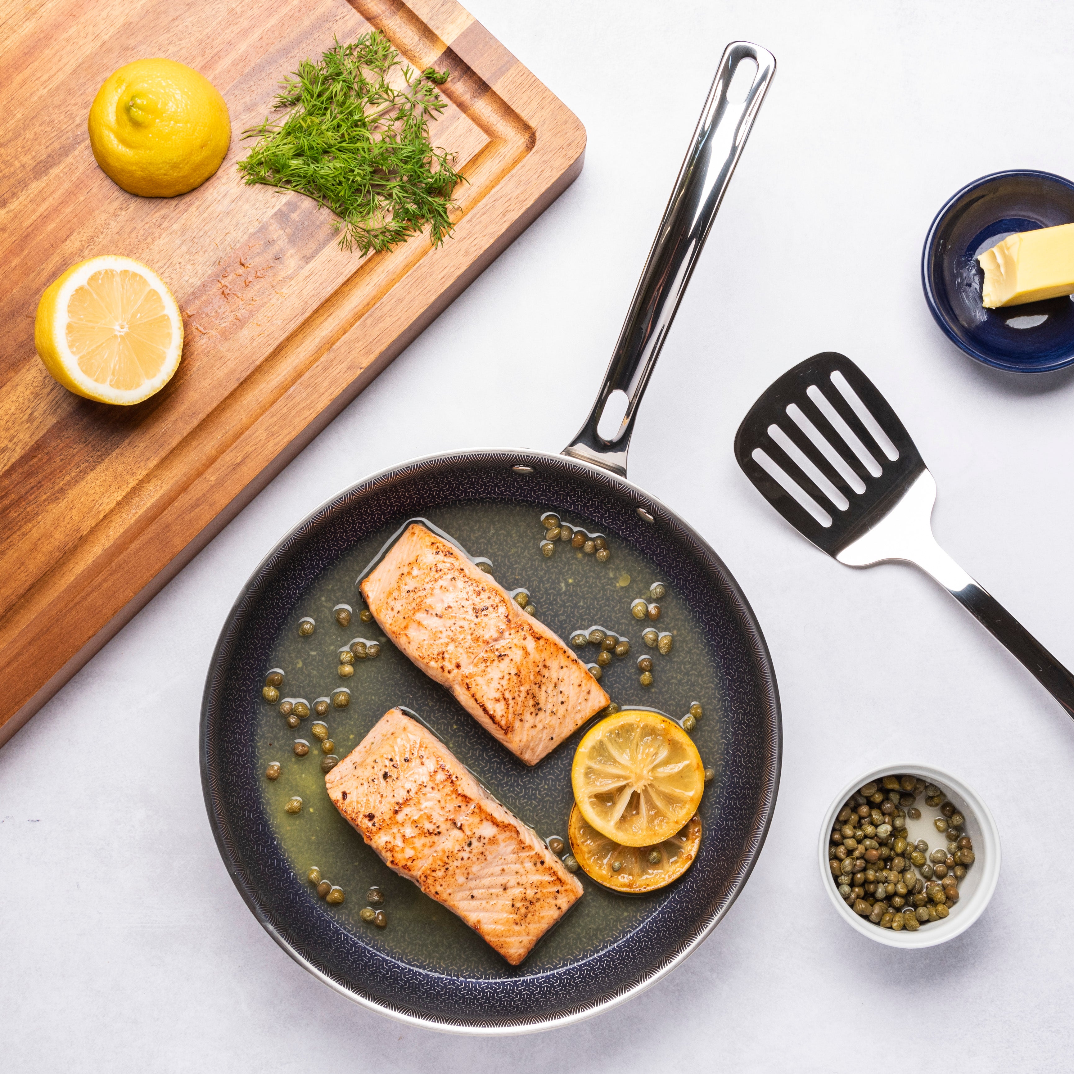 Two cooked salmon fillets with capers and lemon slices in a skillet. Nearby are a spatula, butter in a dish, capers, chopped herbs, a whole lemon, a lemon half, and a cutting board.