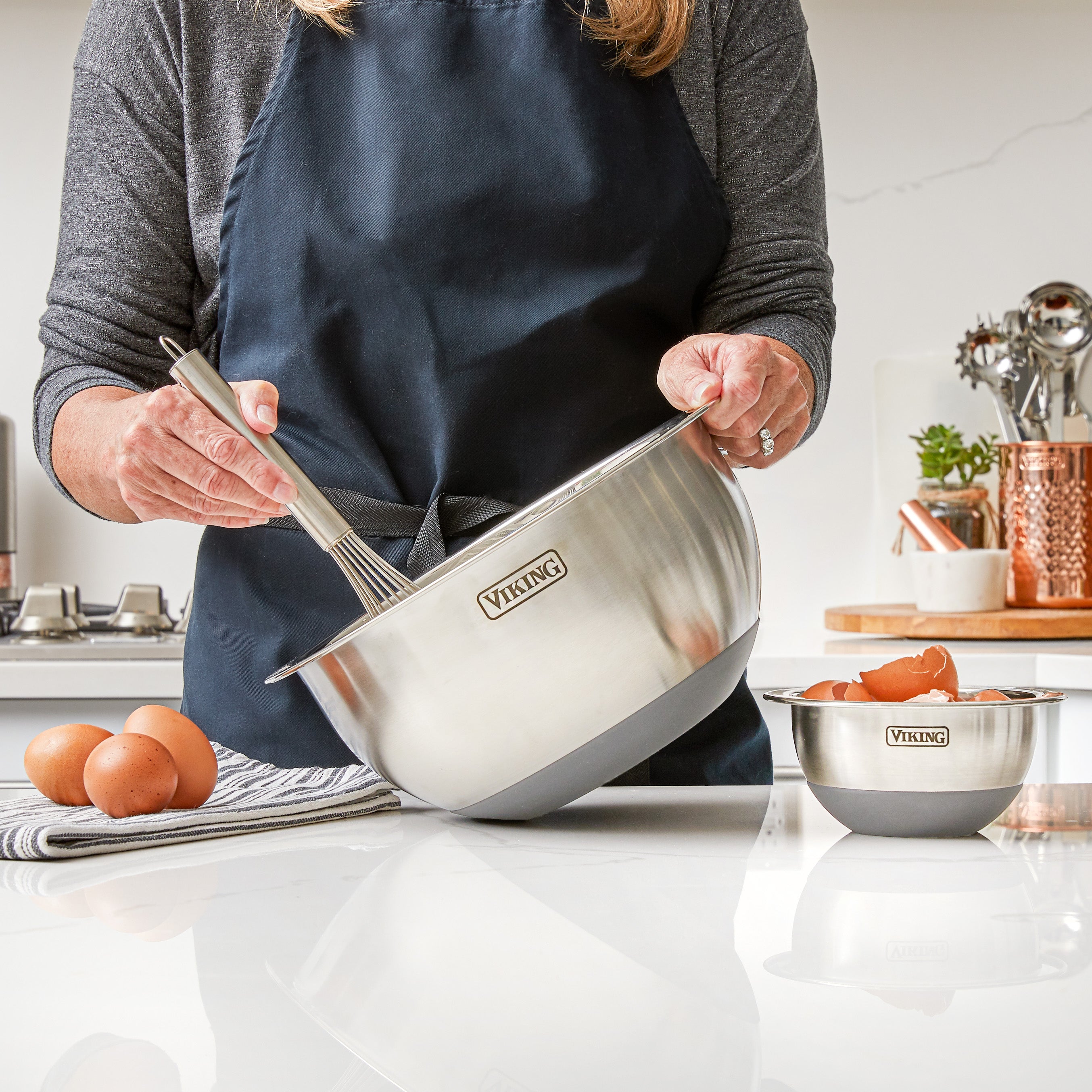 Person in a dark apron whisking ingredients in a large stainless steel Viking bowl, with eggs and another bowl nearby on a white kitchen counter.