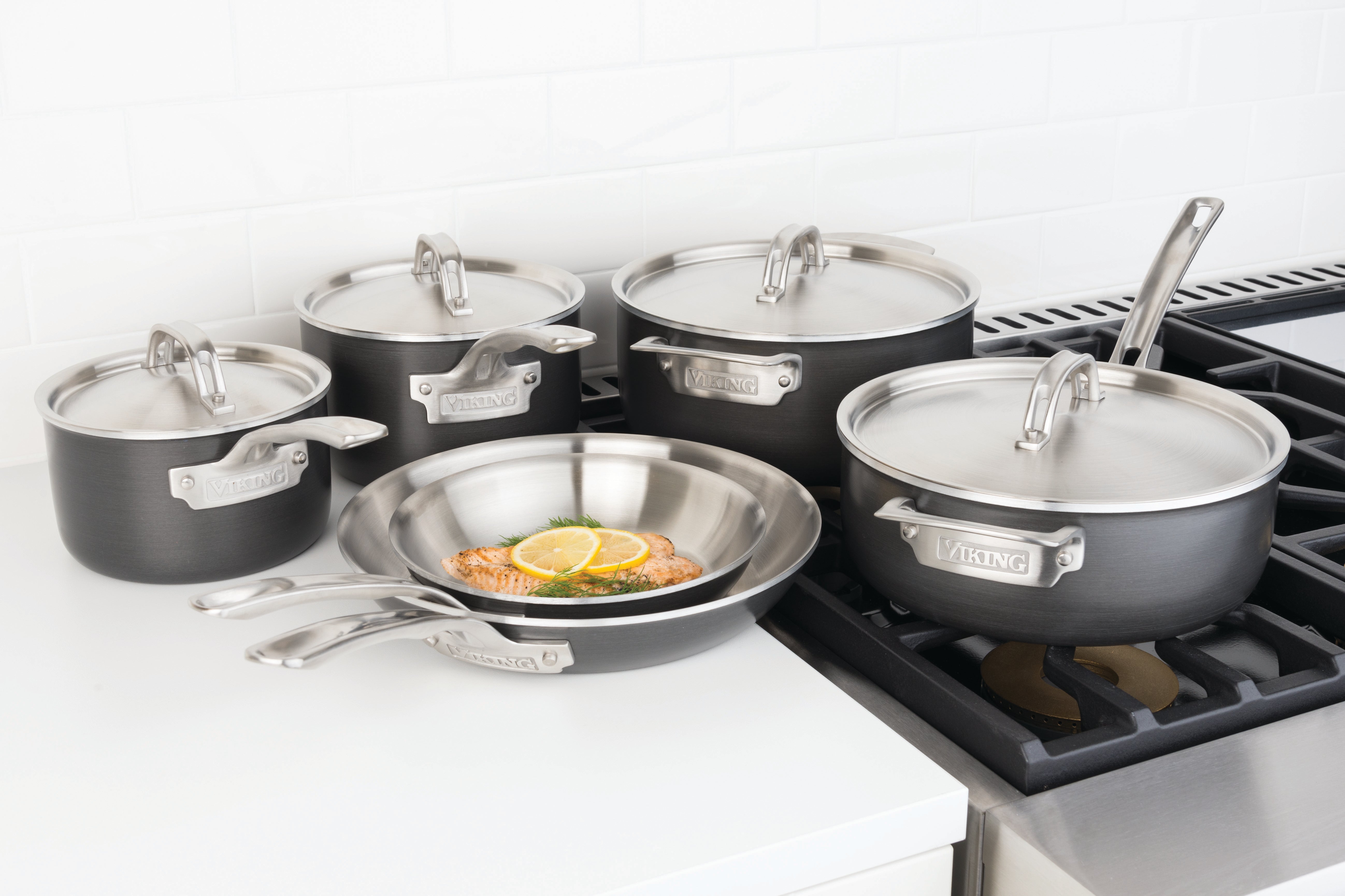 A set of black and stainless steel Viking cookware, including pots with lids and frying pans, is displayed on a white kitchen counter next to a stove. One pan contains cooked salmon with lemon.