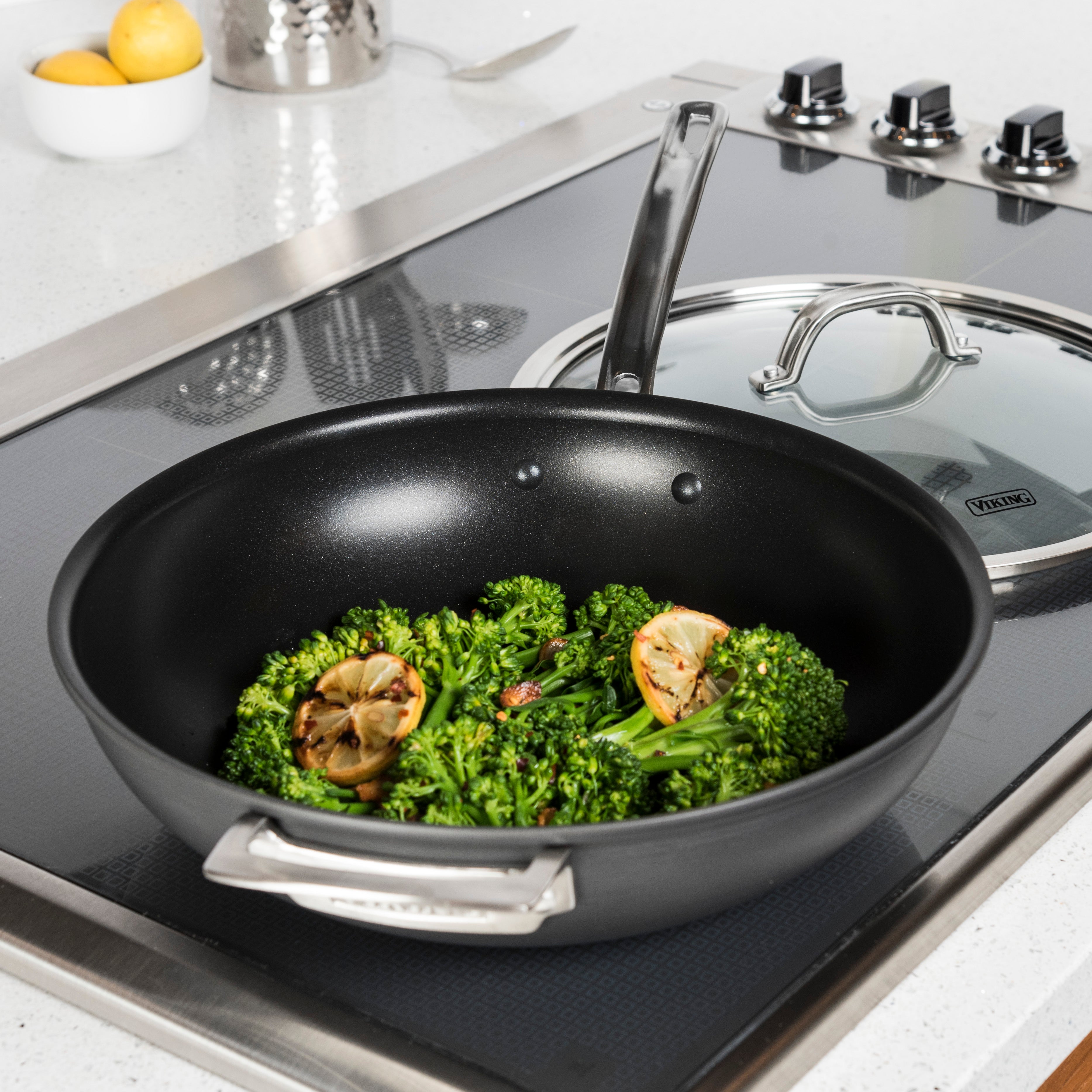 A black nonstick frying pan with sautéed broccolini and charred lemon slices sits on a stove. A glass lid rests nearby on the stovetop, and a white bowl of eggs is in the background.