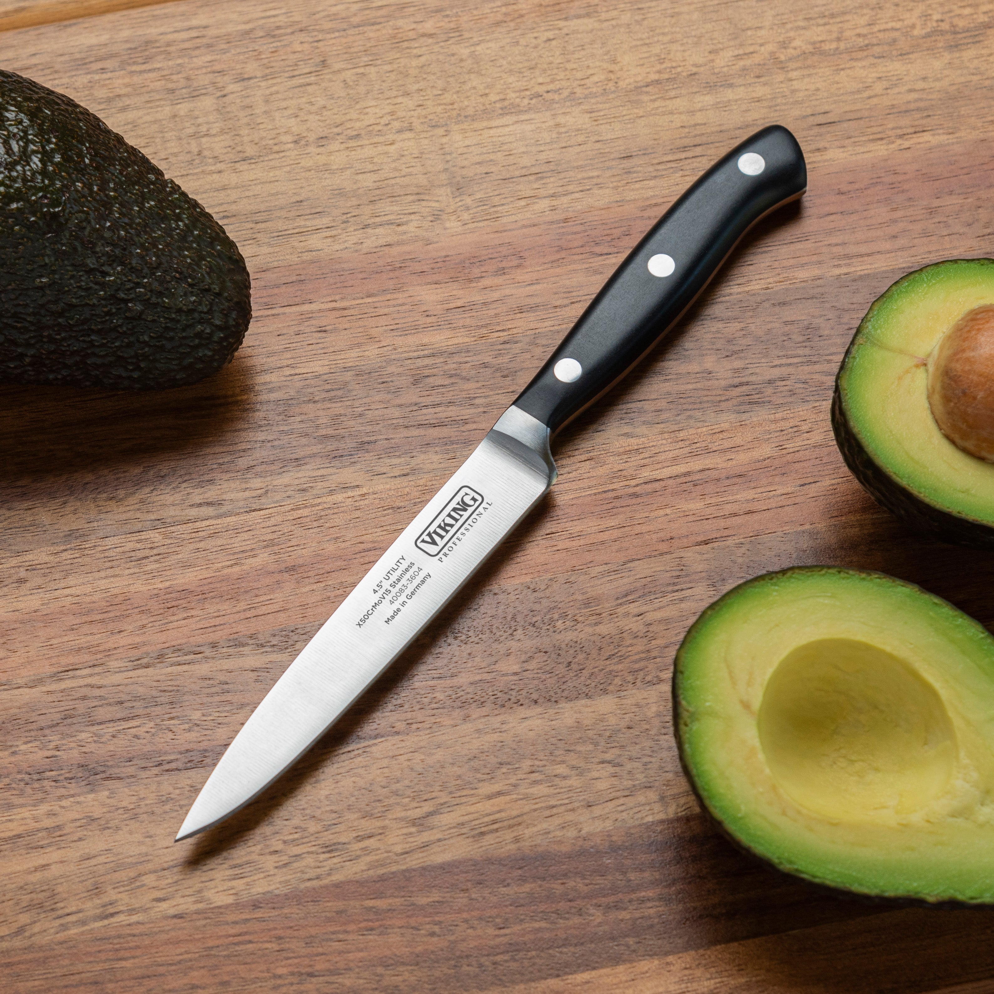 A paring knife with a black handle lies on a wooden cutting board surrounded by a whole avocado, a halved avocado with the pit, and a halved avocado without the pit.
