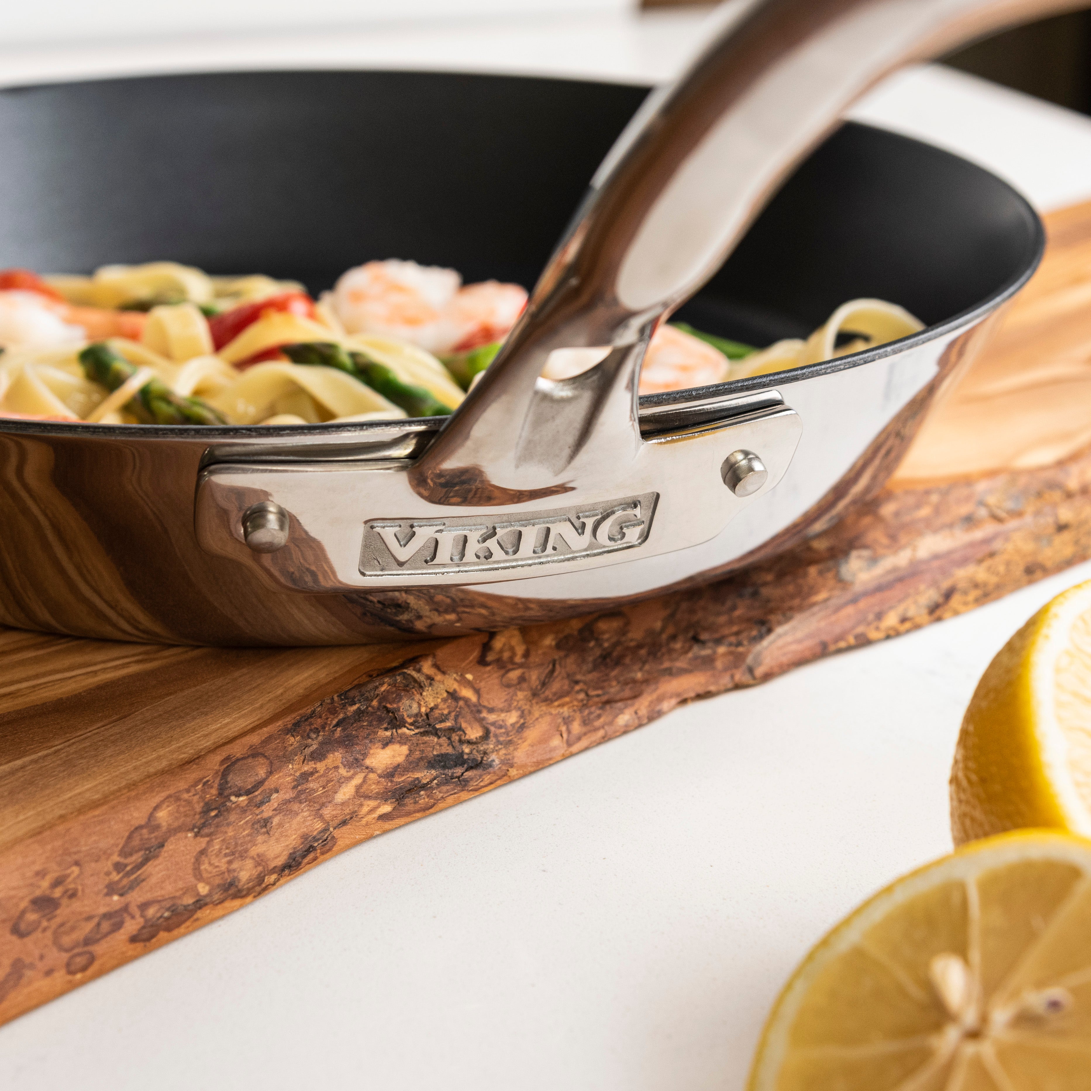 Close-up of a Viking skillet with pasta, vegetables, and shrimp inside, placed on a wooden cutting board with a lemon half in the foreground.