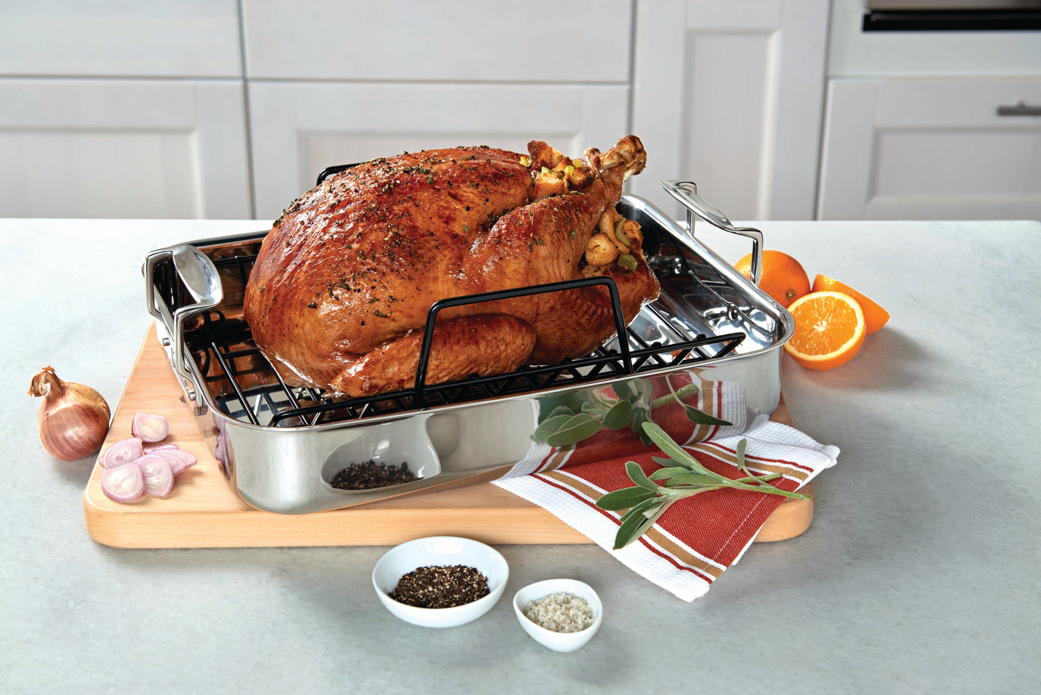 A roasted turkey sits in a metal roasting pan on a wooden cutting board, surrounded by fresh herbs, sliced shallots, orange halves, and small bowls of pepper and salt.