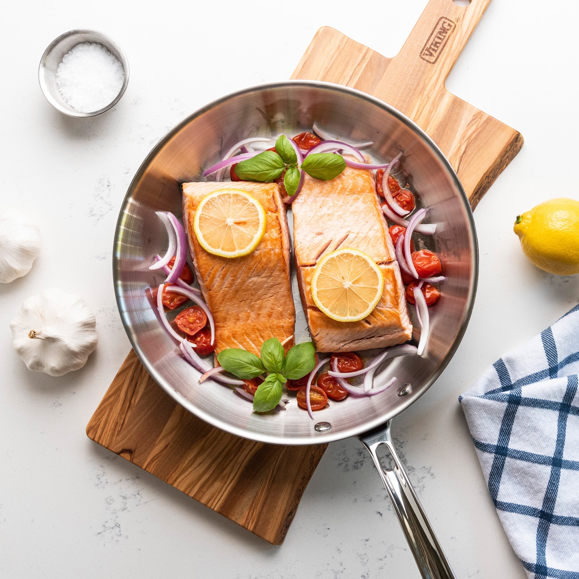 Two salmon fillets in a pan with lemon slices, cherry tomatoes, red onion, and basil. Surrounded by garlic, salt, a lemon, and a blue-checked towel on a white countertop.