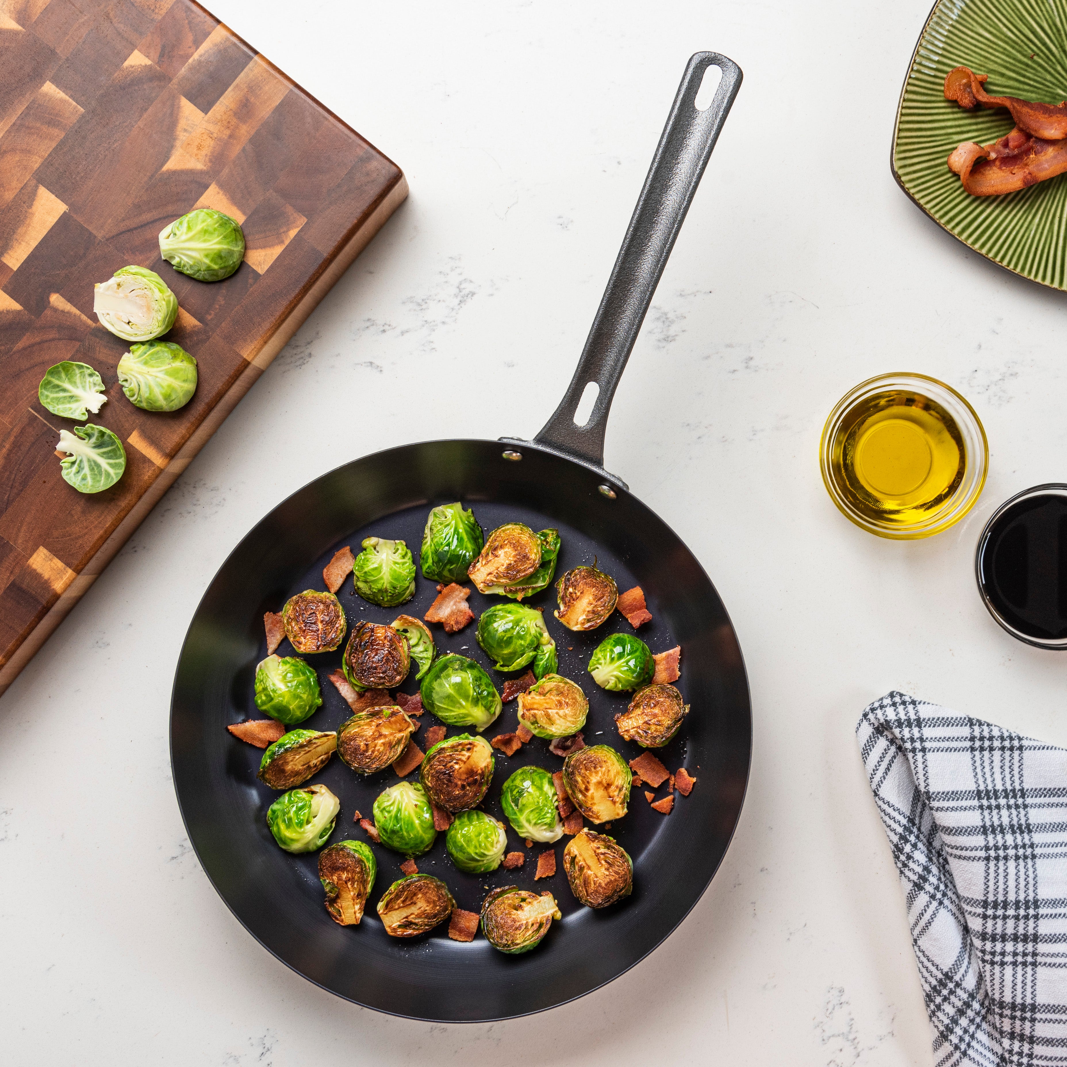 A frying pan with cooked Brussels sprouts and bacon sits on a white counter, surrounded by a cutting board, sliced Brussels sprouts, olive oil, a plaid towel, and a green plate with bacon.