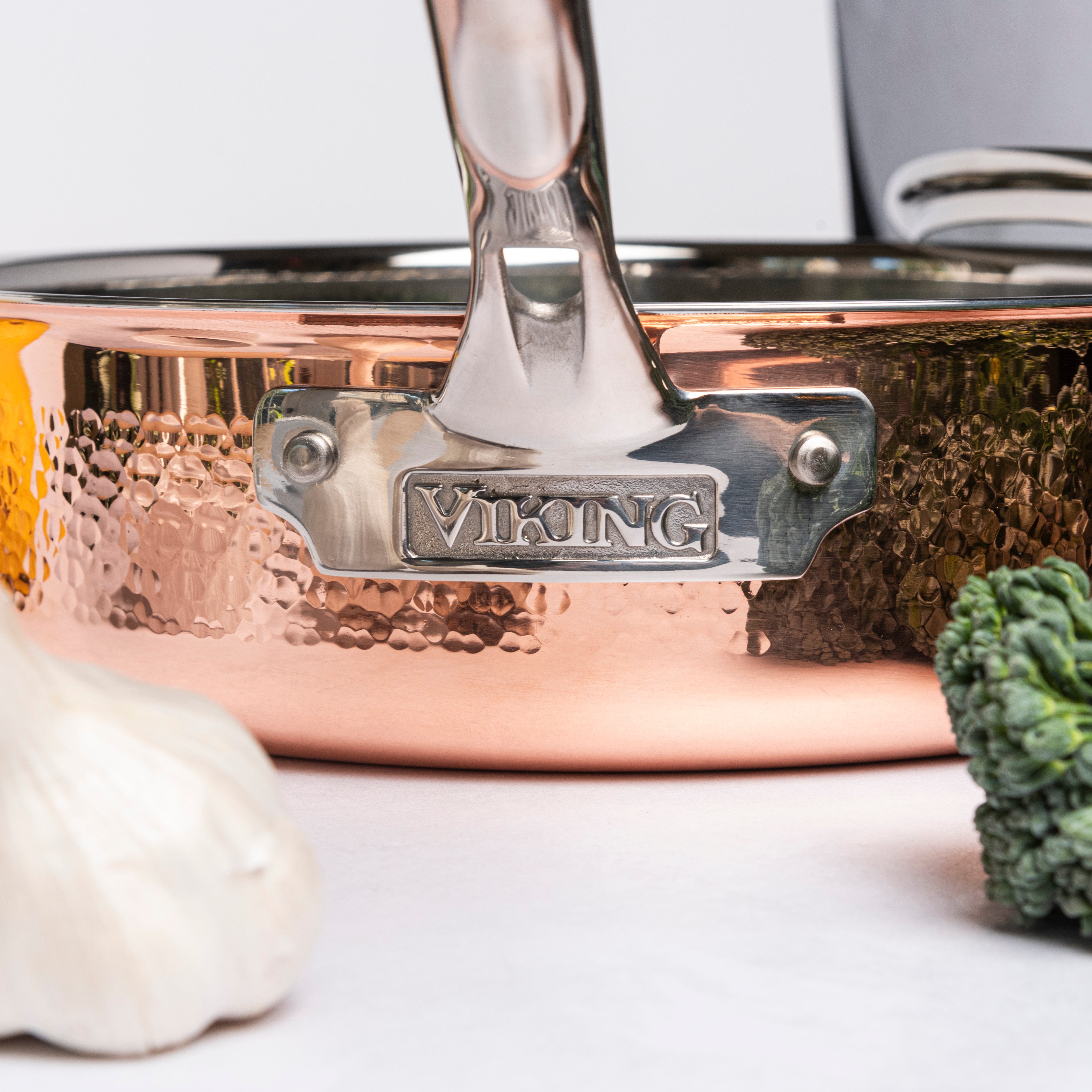 A close-up of a Viking copper saucepan with a shiny handle, surrounded by fresh garlic and broccoli on a white surface.
