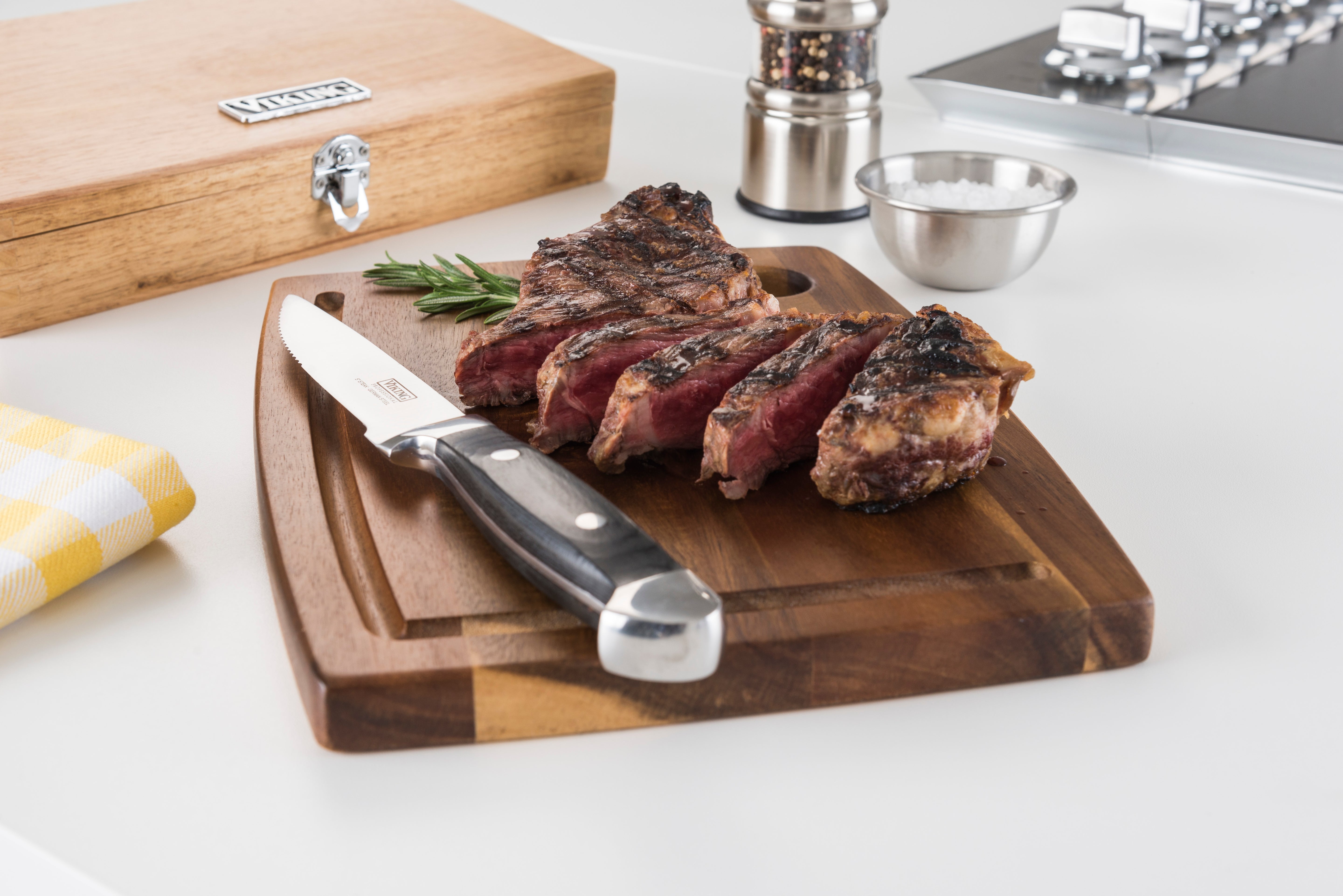 Sliced grilled steak on a wooden cutting board with a knife, garnished with rosemary, next to a container of salt, pepper grinder, and a wooden box on a white kitchen counter.