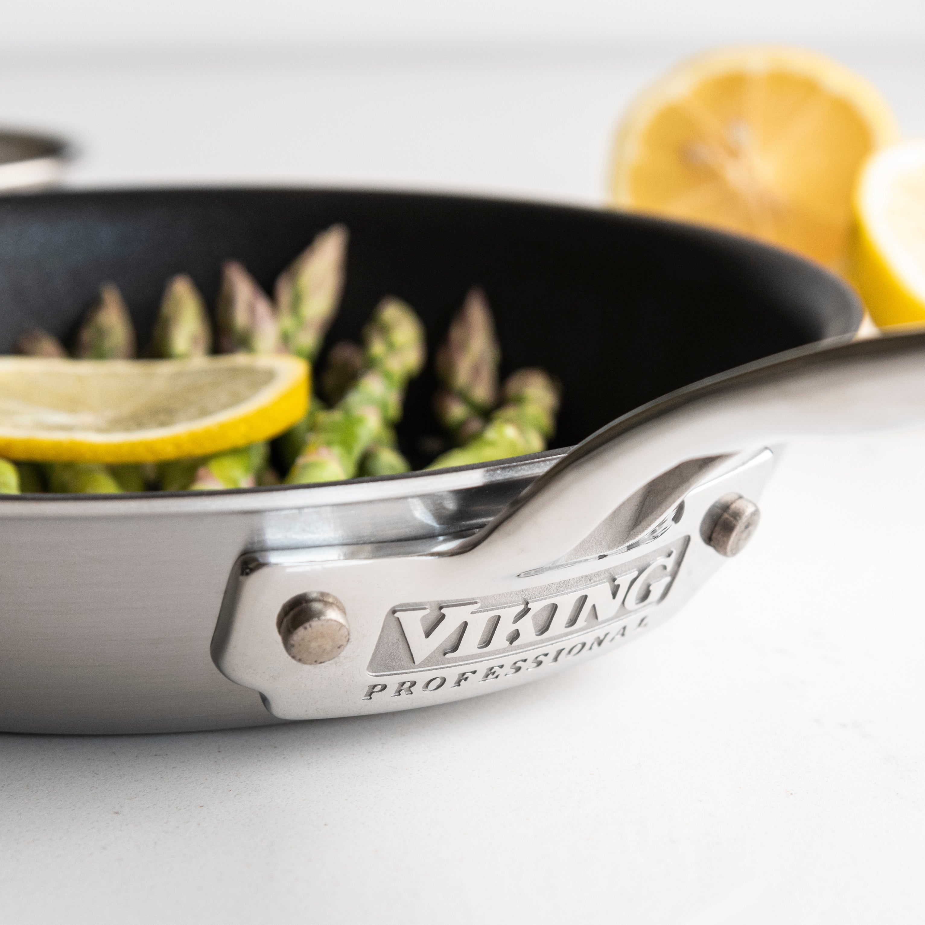 A stainless steel Viking Professional pan with asparagus spears and a lemon slice inside. A halved lemon is blurred in the background on a white surface.