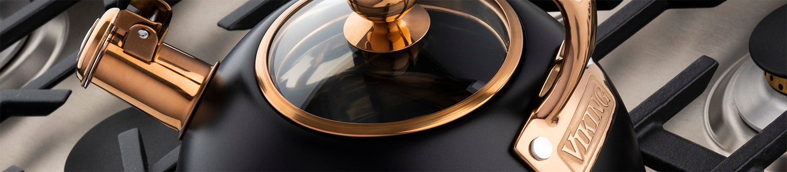 A close-up of a black and copper kettle with a glass lid, sitting on a modern gas stovetop with metal grates and burners visible in the background.
