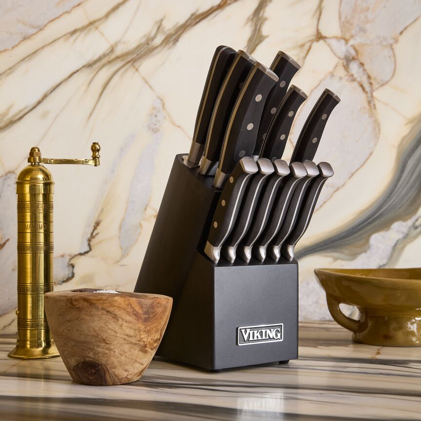 A Viking Culinary Products 14-Piece X50CrMoV15 Steel Cutlery Set with a black block sits on a striped countertop, accompanied by a wooden bowl, a brass pepper grinder, and a green ceramic dish, all set against a marble-patterned backsplash.