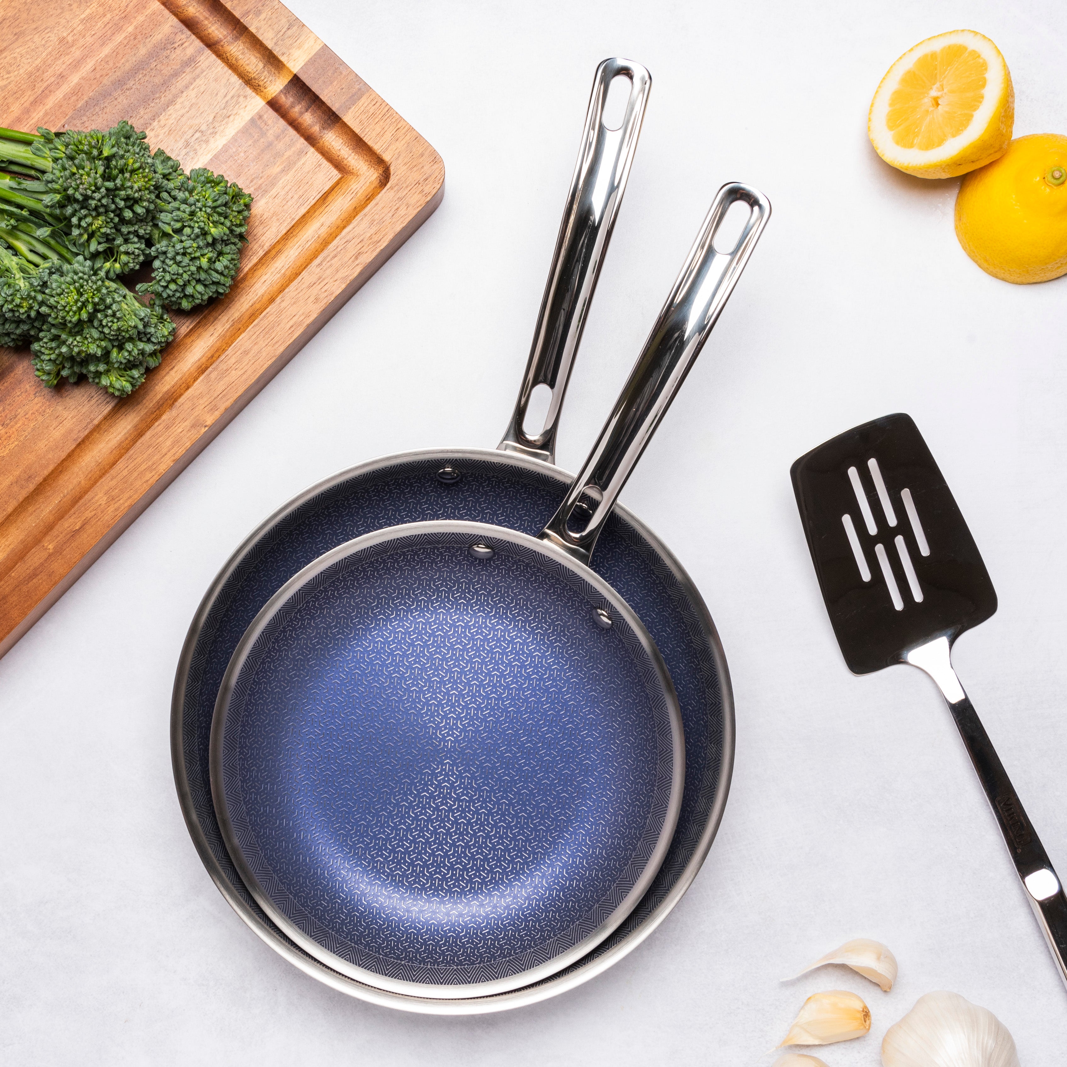 The Viking 3-Ply Hybrid Plus Ceramic 2 Pc Nonstick Fry Pan Set by Viking Culinary Products (9.5" & 11") nests together on a white surface beside a wooden cutting board topped with broccolini, lemon halves, peeled garlic cloves, and a spatula.