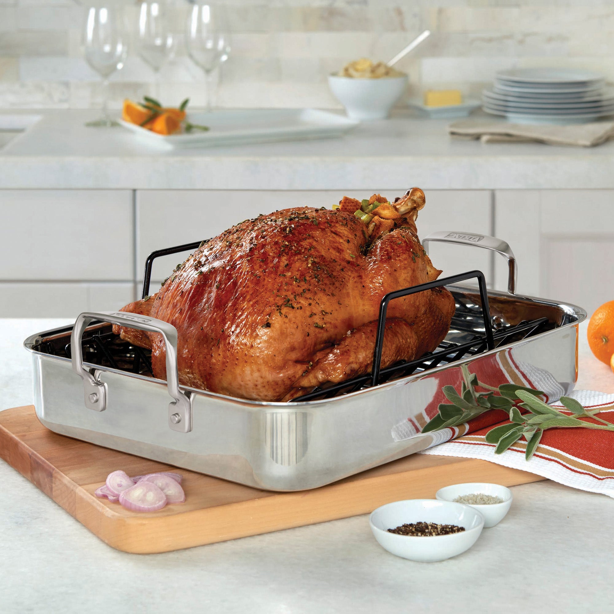 A Viking 3-Ply Stainless Steel Roaster with Rack by Viking Culinary Products sits on the counter, filled with roasted turkey and herbs. A bonus digital thermometer, plates, bowls of spices, and utensils are arranged nearby.