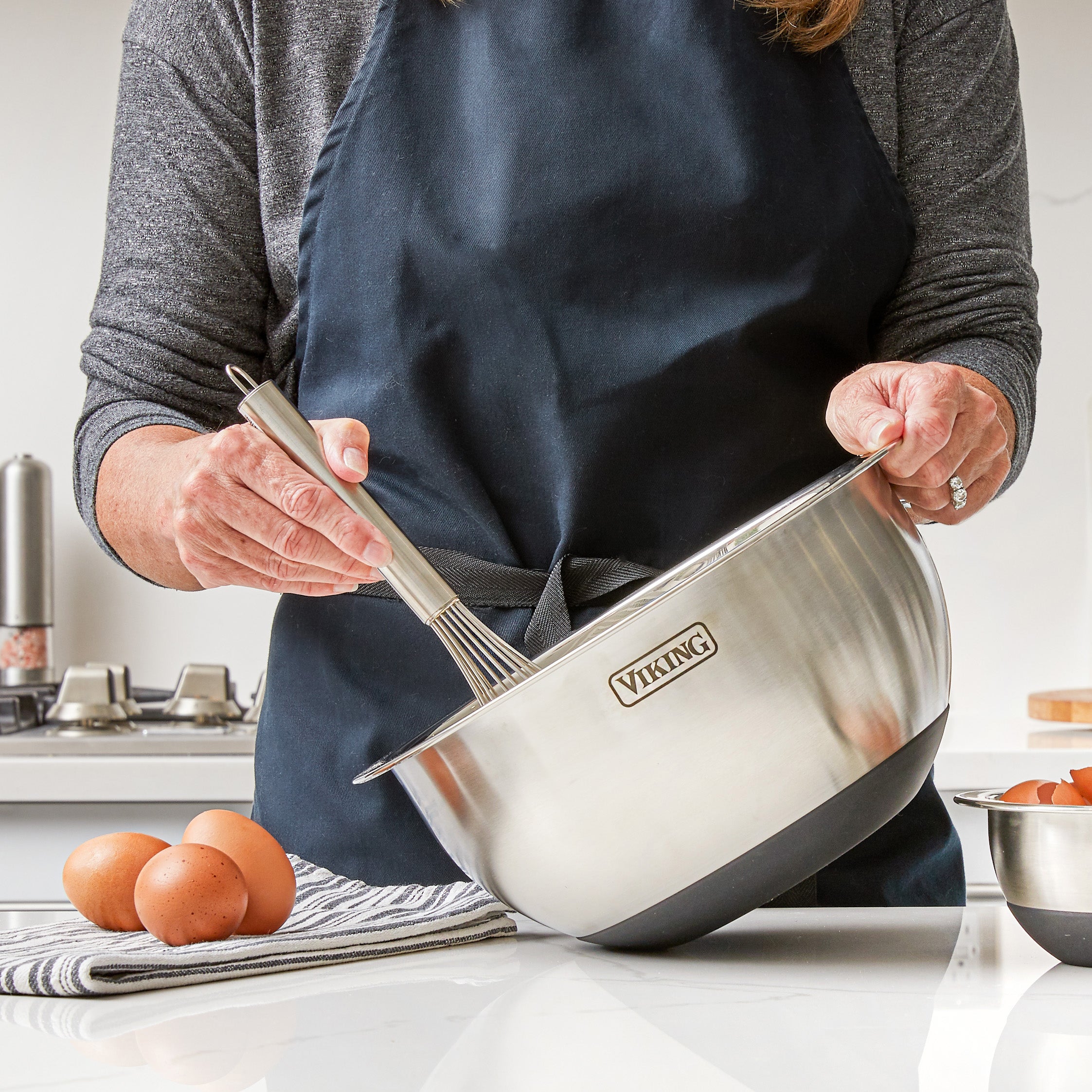A person in a dark apron holds a Viking 9-Piece Stainless Steel Mixing Bowl with a whisk. Three brown eggs sit on a striped towel, and another Viking bowl with airtight lids is nearby on the kitchen counter.