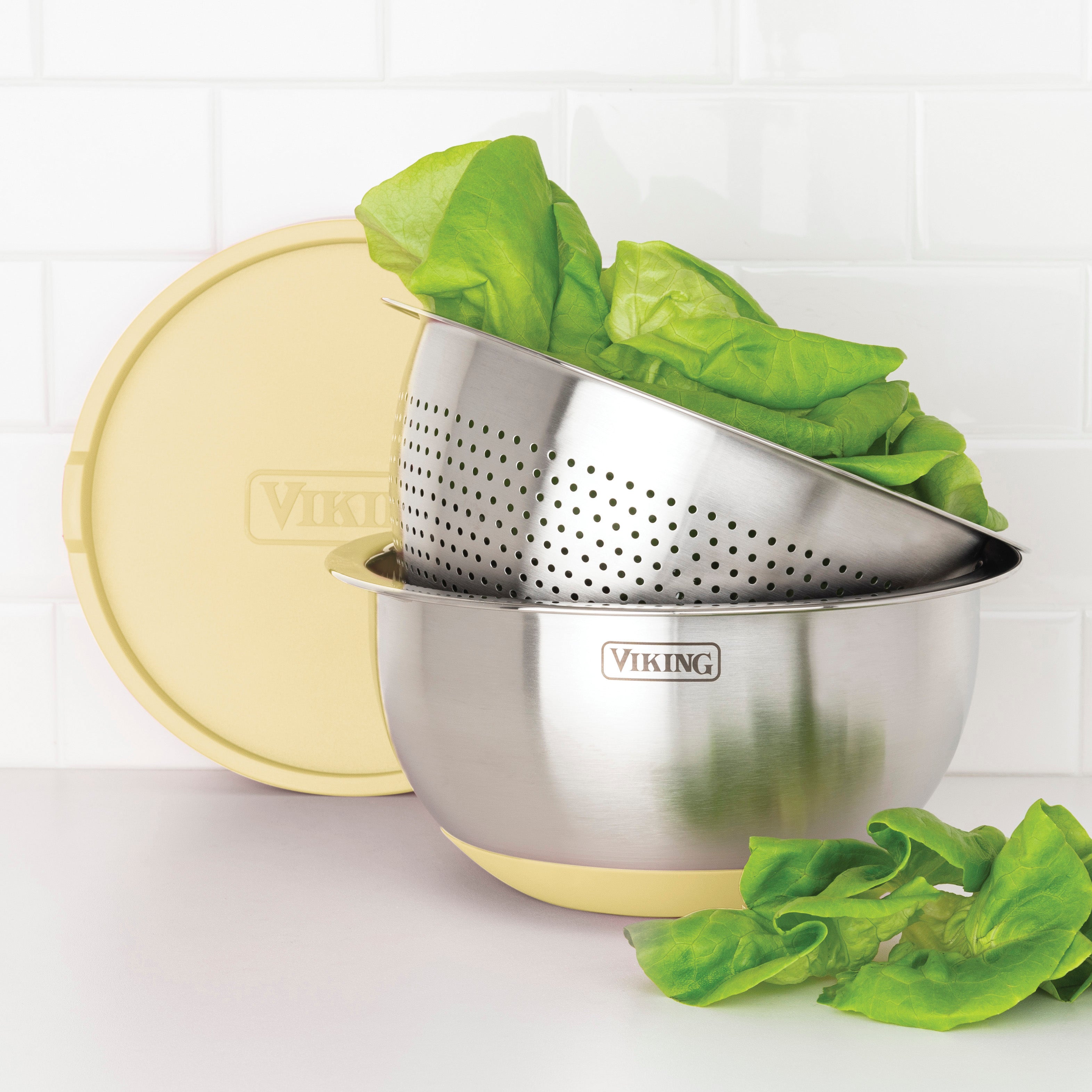 A Viking Culinary 9-Piece Stainless Steel Mixing Bowl Set with Strainer in Buttermilk is shown, filled with leafy lettuce. A yellow lid rests nearby, while extra lettuce leaves are scattered in front of the bowls.