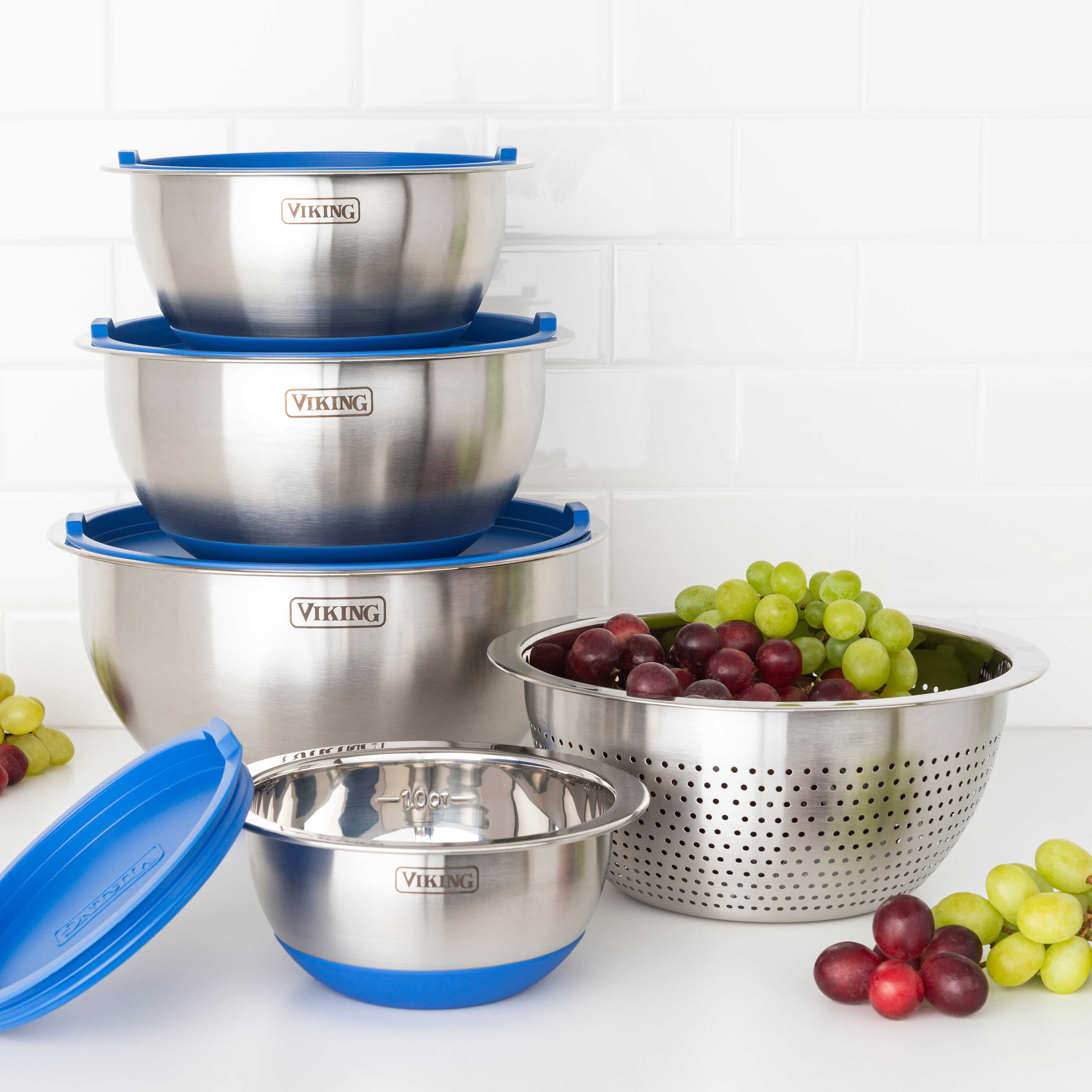 The Viking Culinary Products 9-Piece Stainless Steel Mixing Bowl Set with Strainer in Blue, featuring airtight lids and stackable bowls for compact storage, sits beside a grape-filled strainer on a white countertop against a tiled backsplash.