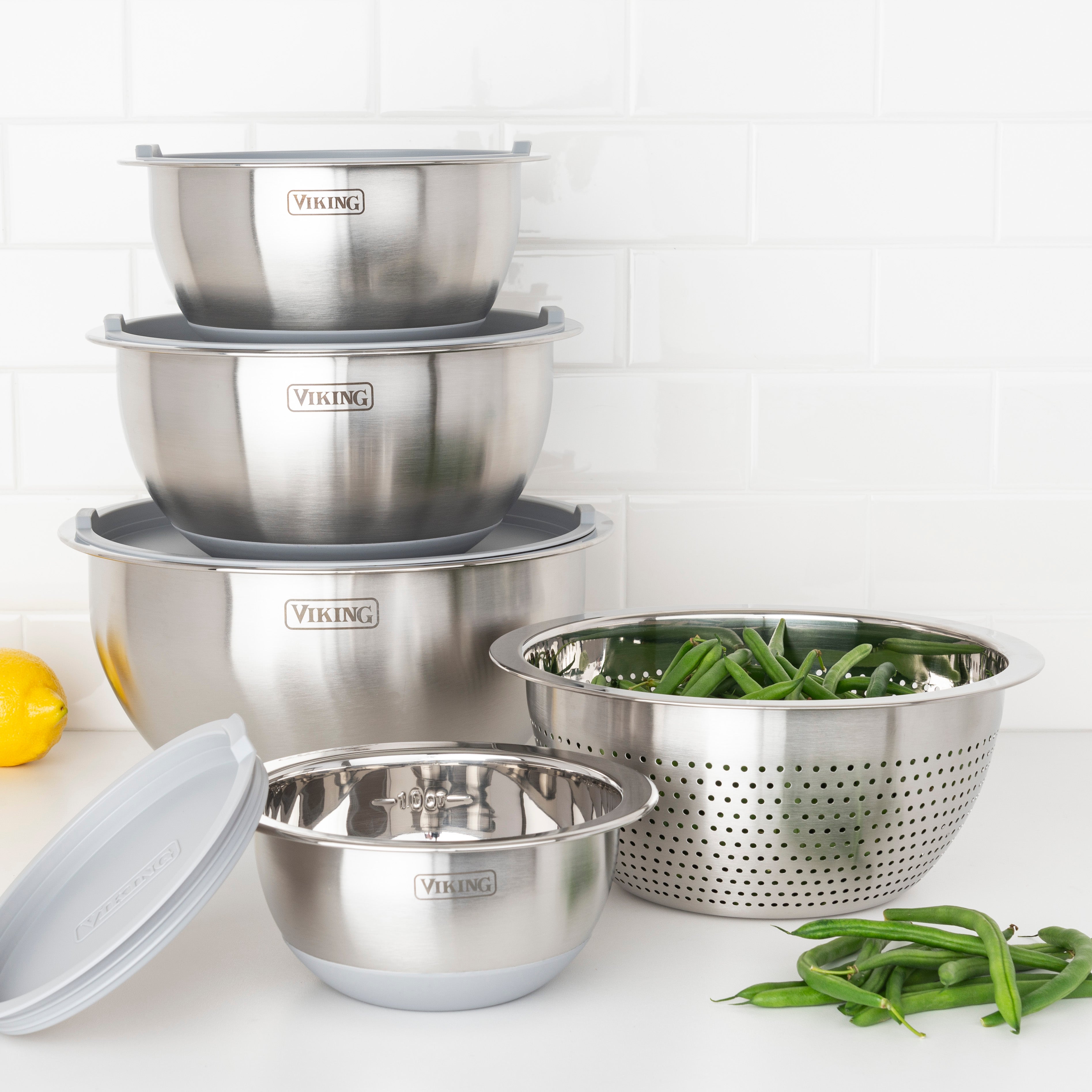 The Viking 9-Piece Stainless Steel Mixing Bowl Set with Strainer by Viking Culinary Products is stacked on a kitchen counter, with a metal strainer filled with green beans and a lemon nearby, all set against a white tile backsplash.