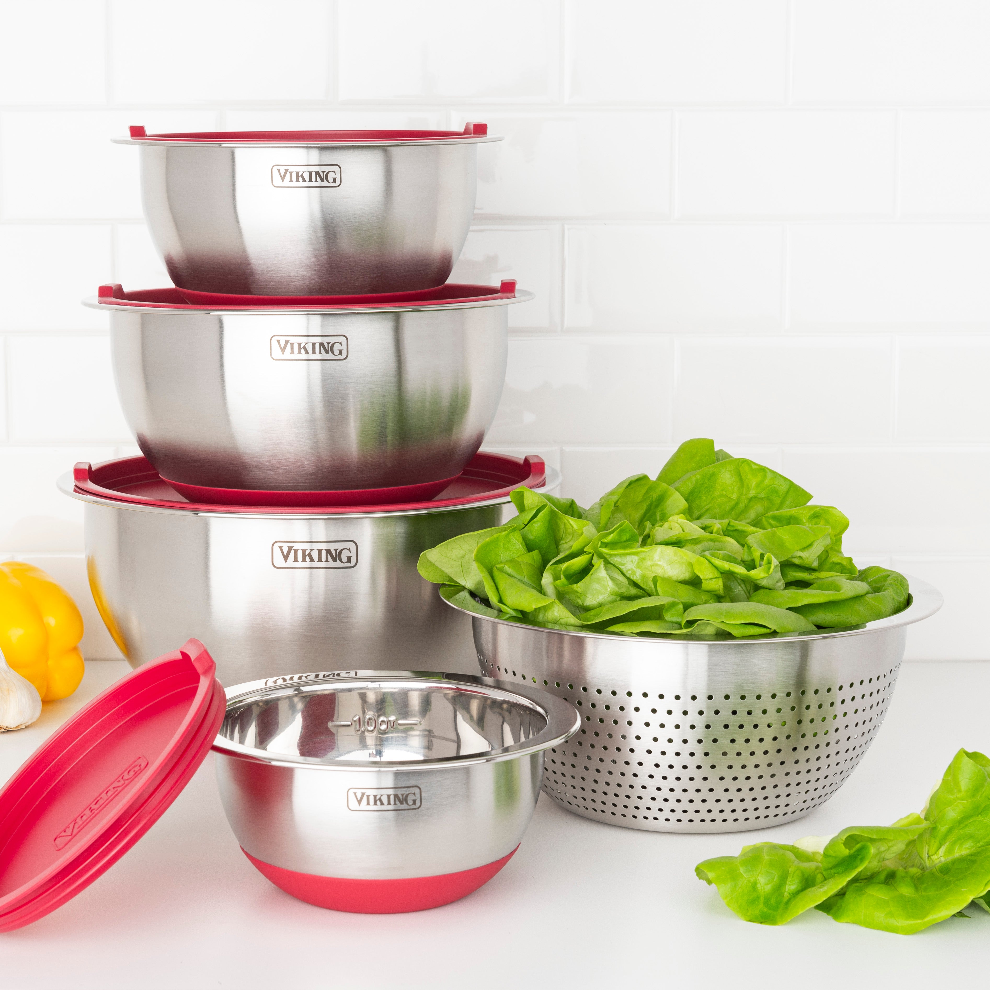 The Viking 9-Piece Stainless Steel Mixing Bowl Set with Strainer by Viking Culinary Products, featuring red accents and airtight lids, is stacked compactly beside a colander of spinach on a white countertop with a tile backsplash.