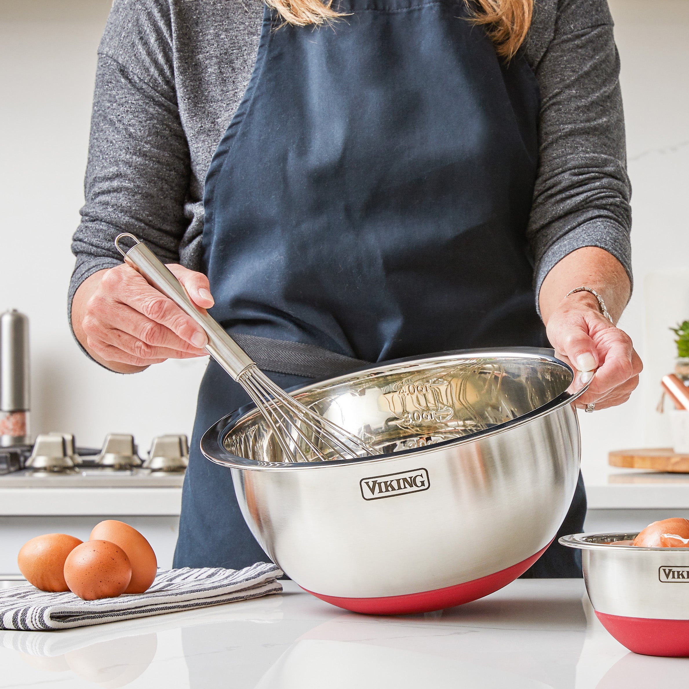 A person in a dark apron uses a whisk in a Viking 9-Piece Stainless Steel Mixing Bowl Set with Strainer by Viking Culinary Products on the counter, with two eggs, a striped towel, and another bowl nearby; airtight lids allow compact storage.