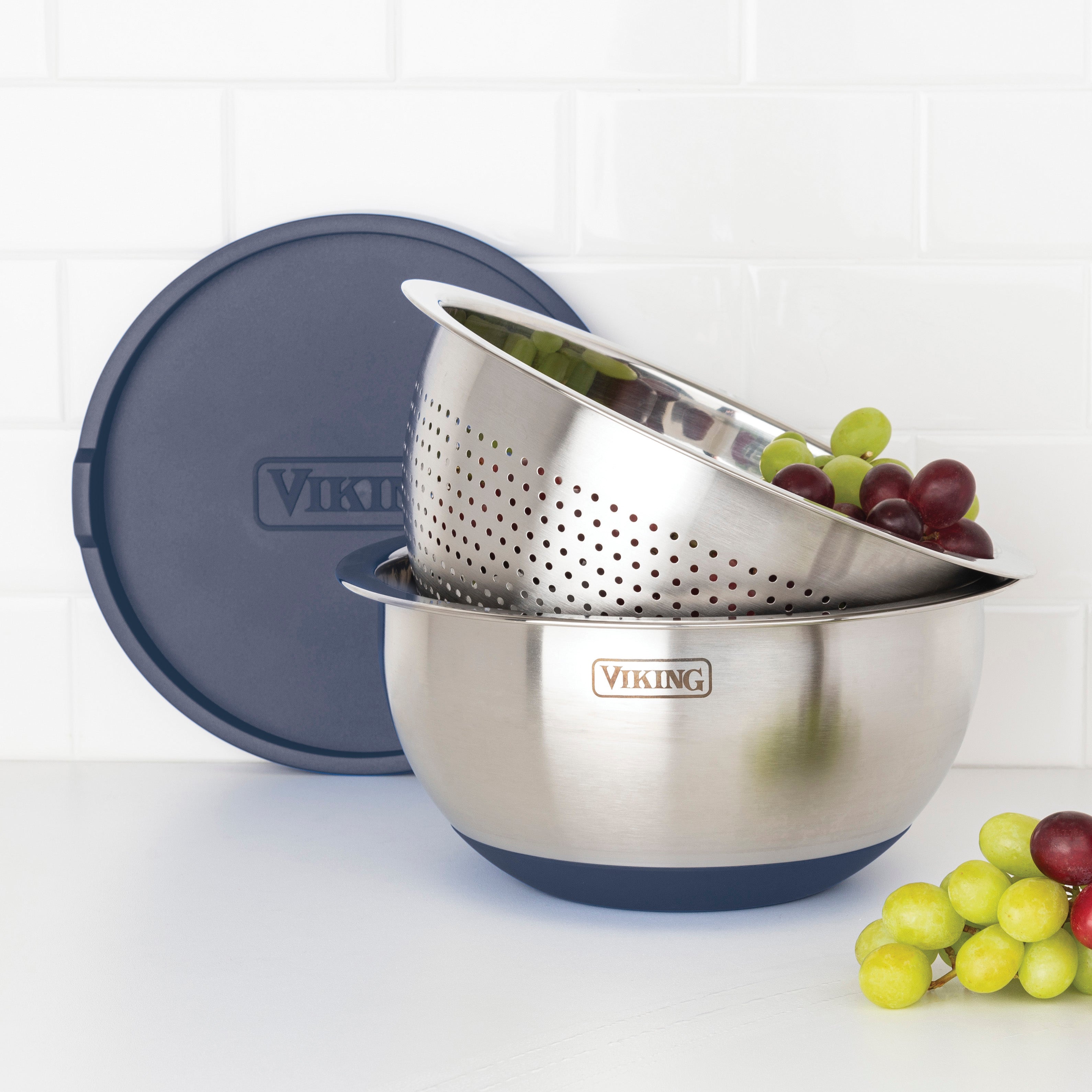 The Viking 9-Piece Stainless Steel Mixing Bowl Set with Strainer by Viking Culinary Products features slate blue lids, a mixing bowl, and a perforated colander, displayed on a white surface in front of a tiled wall.