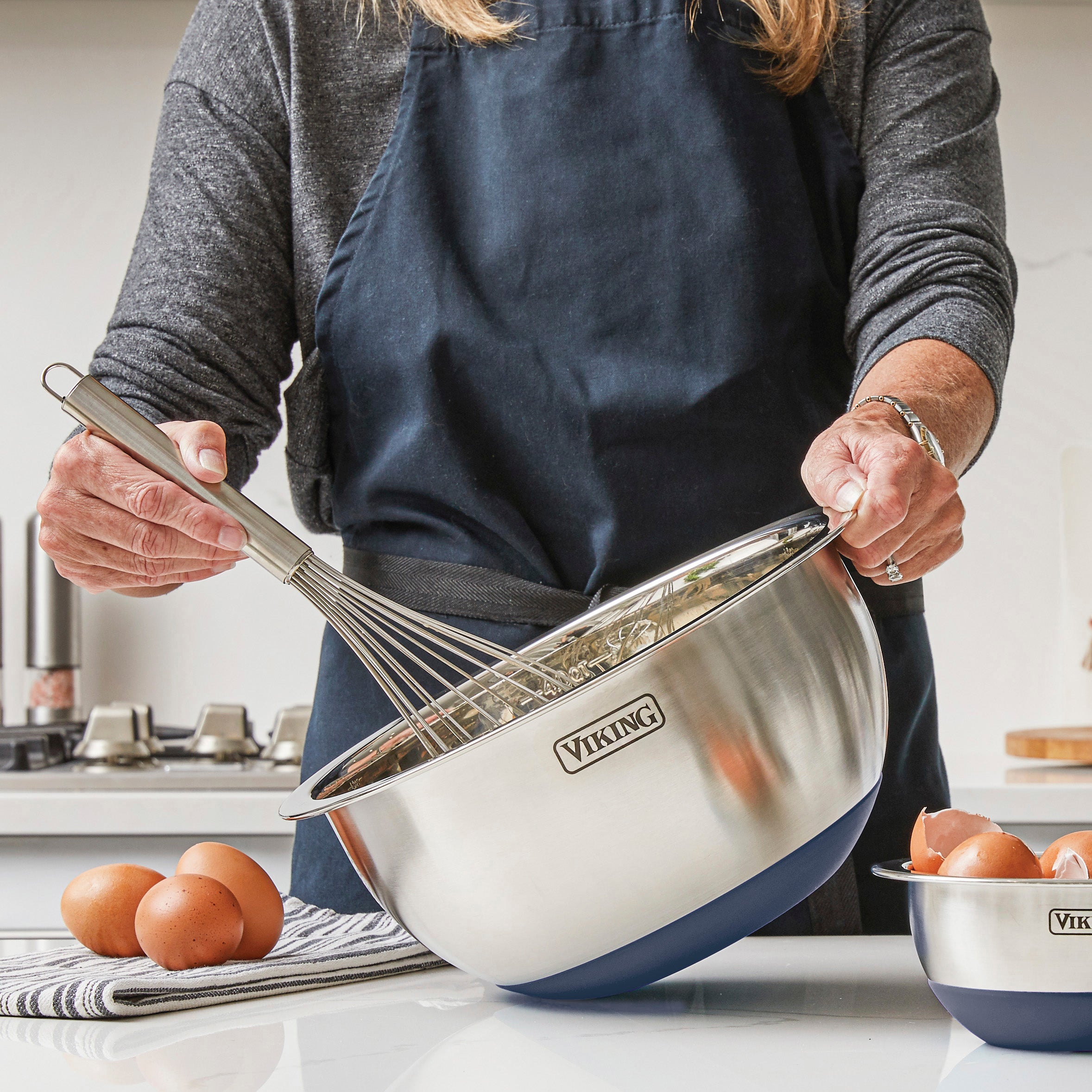 Wearing a dark apron, a person whisks ingredients in a Viking Culinary Products 9-Piece Stainless Steel Mixing Bowl with Strainer, Slate Blue, eggs on a striped towel and another bowl nearby on the kitchen counter.