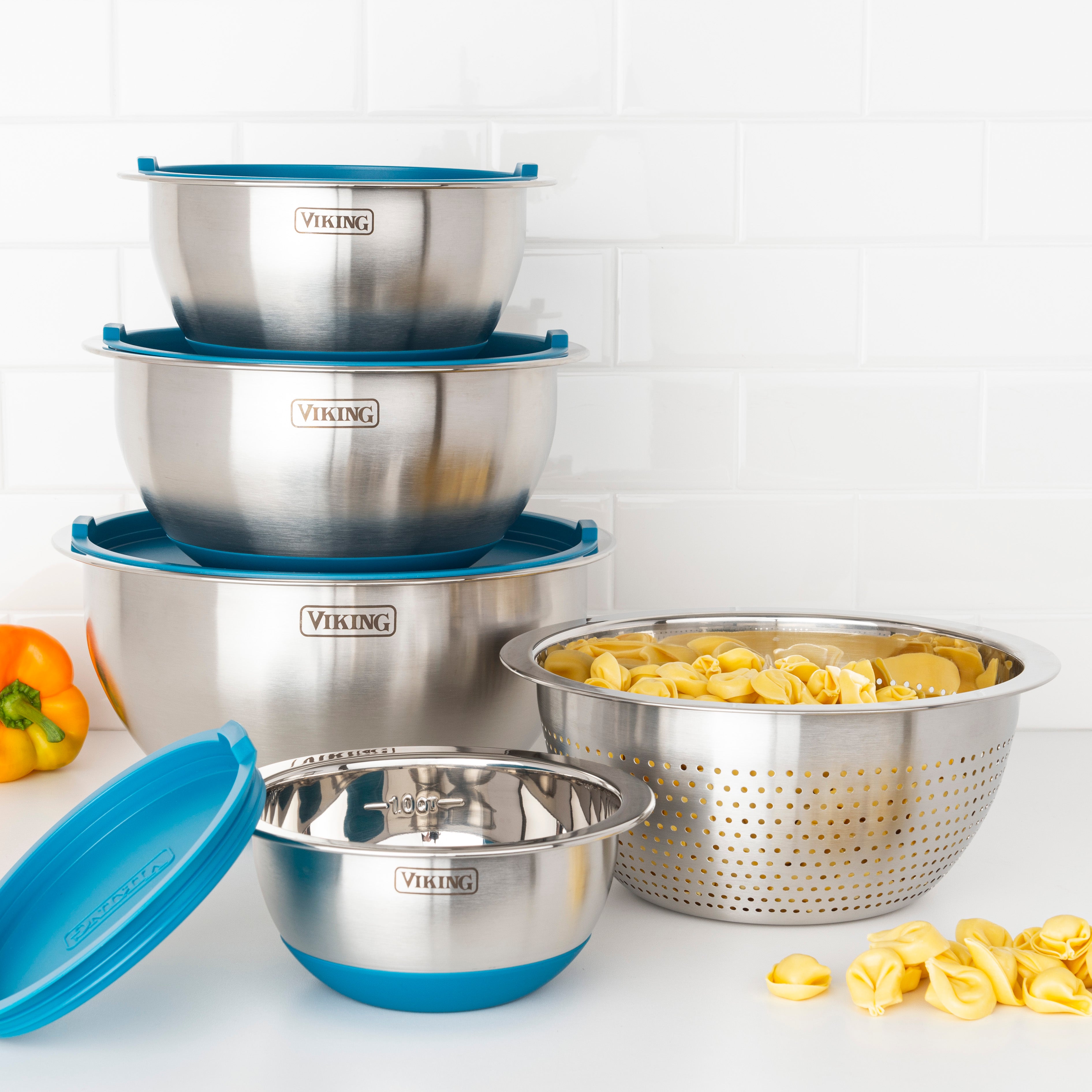 The Viking 9-Piece Stainless Steel Mixing Bowl Set with Strainer by Viking Culinary Products features teal-accented bowls and airtight lids, neatly stacked beside a colander of tortellini on a white counter, with a yellow bell pepper and extra lid nearby.