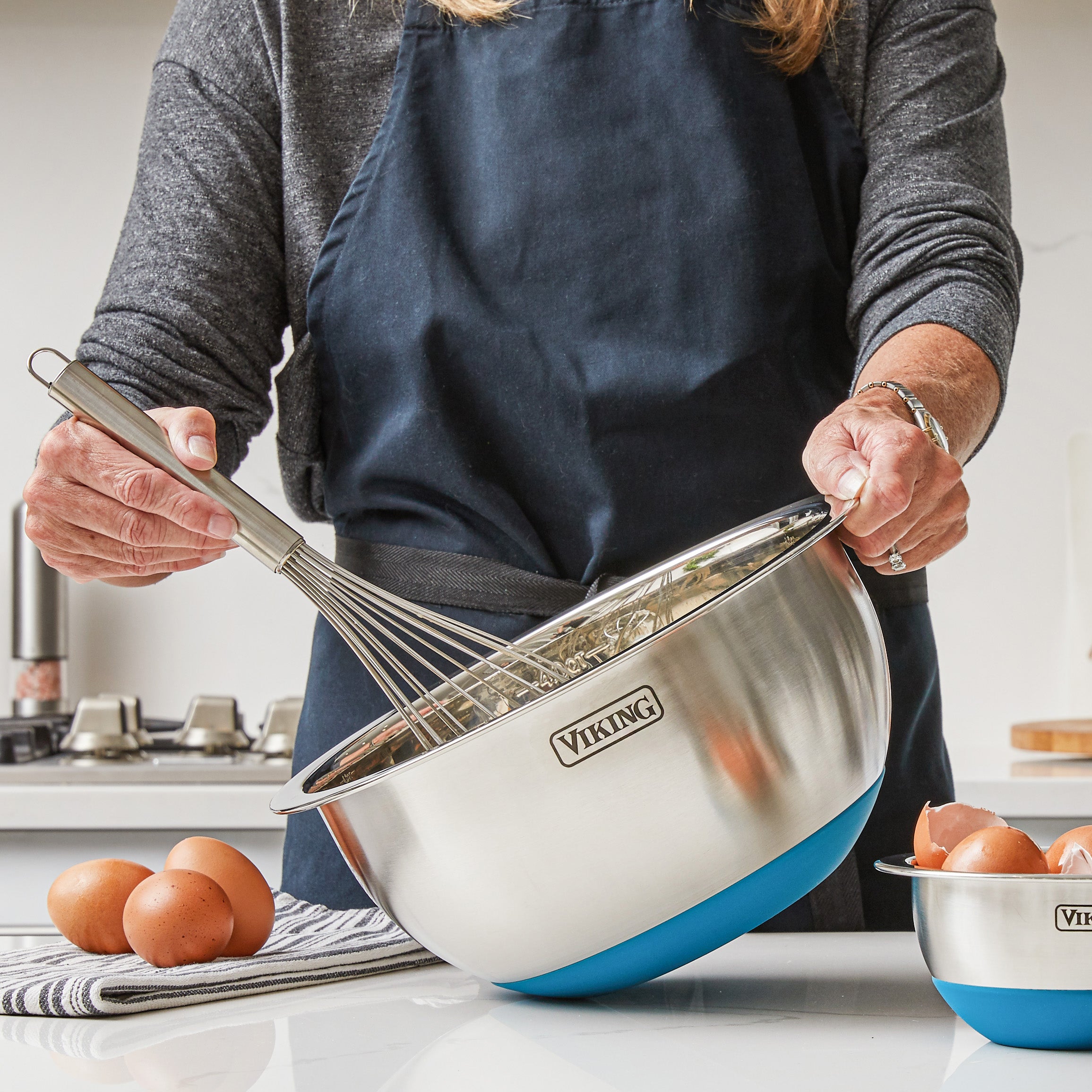 A person in a dark apron uses a whisk in a large Viking 9-Piece Stainless Steel Mixing Bowl from Viking Culinary Products. Brown eggs, striped cloth, and smaller bowls are nearby, highlighting compact storage in a modern kitchen.