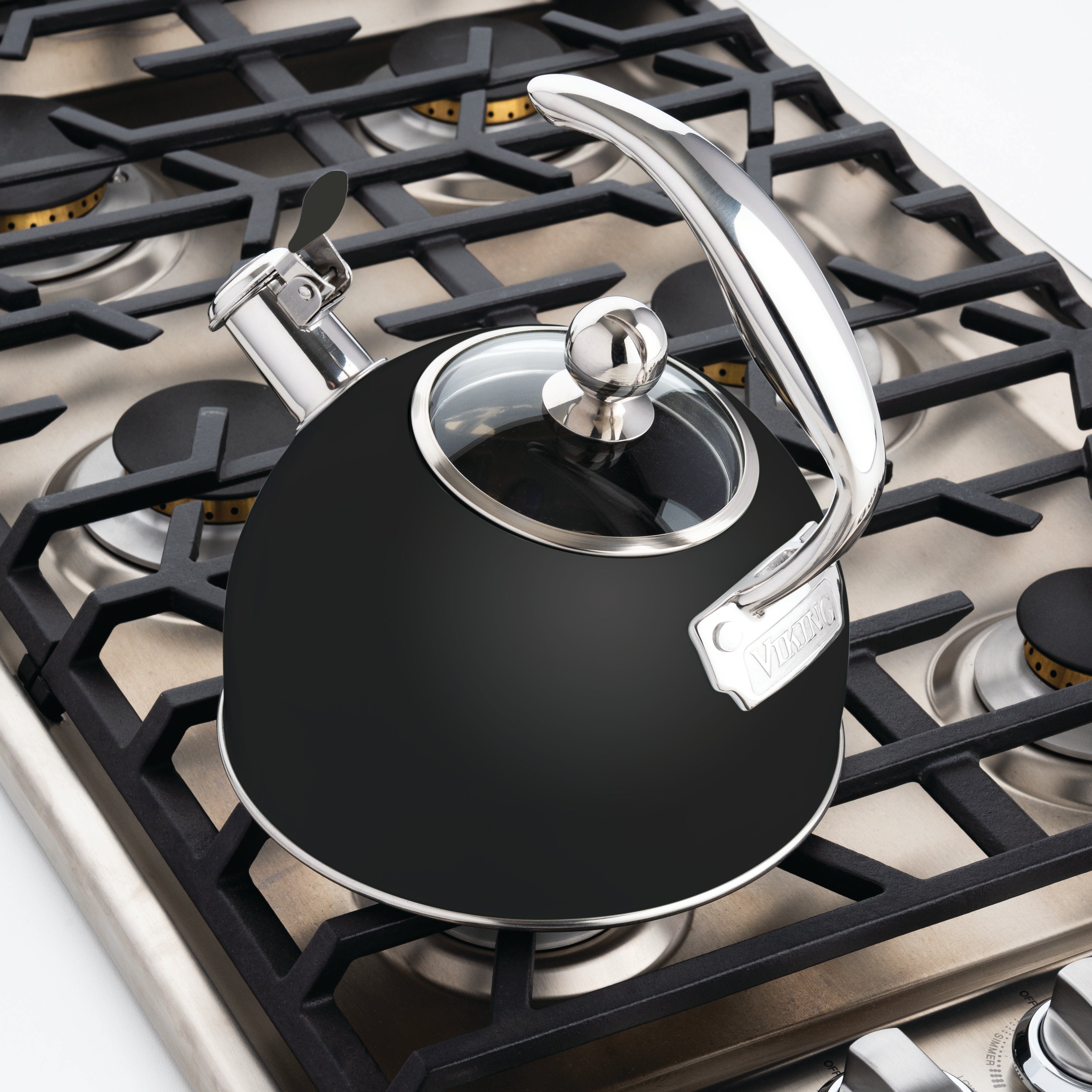 A Viking 2.6 Qt. Stainless Steel Whistling Kettle with a matte black finish, from Viking Culinary Products, sits on a four-burner gas stove with cast iron grates. The kettle’s whistling spout signals when water boils.