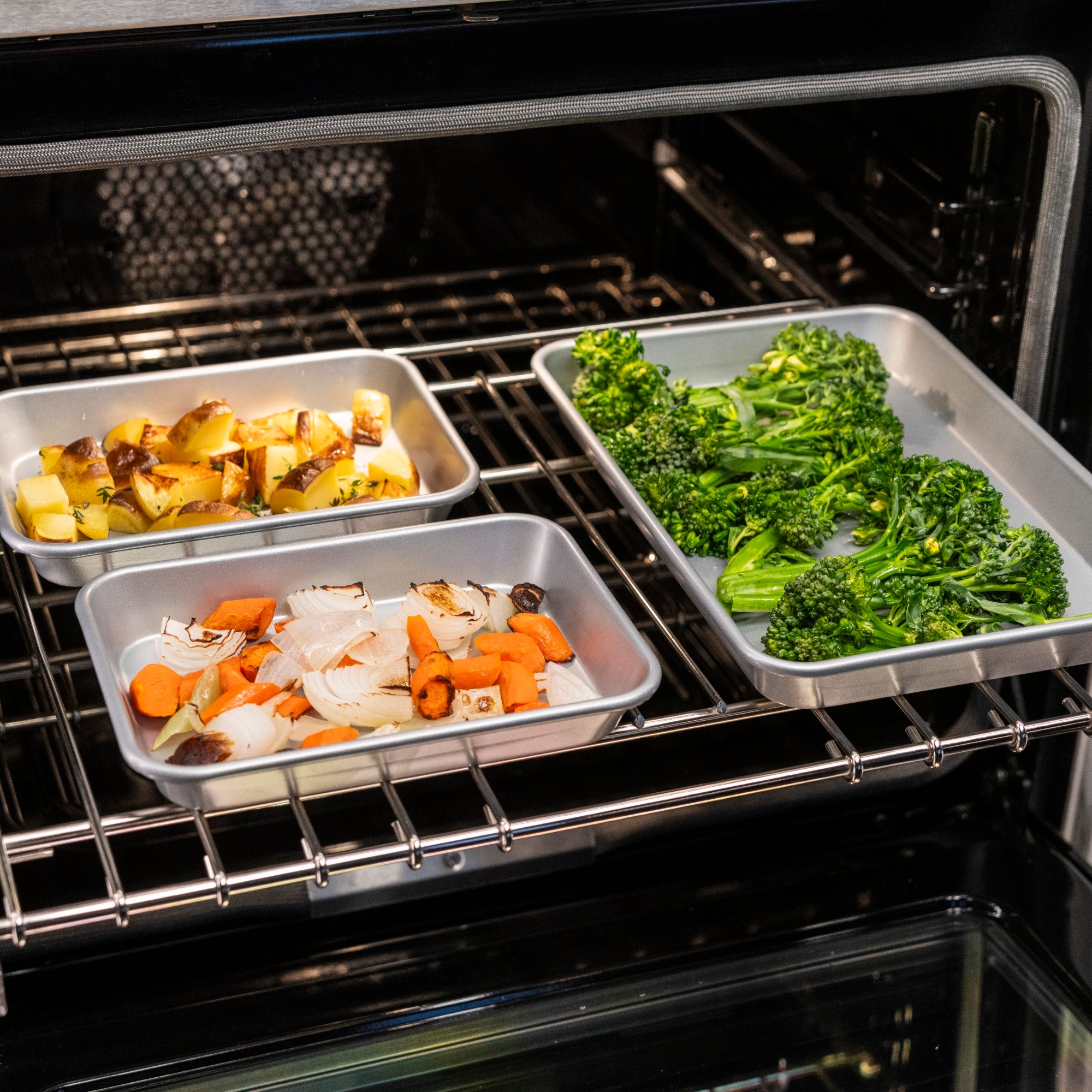 Three trays from the Viking Aluminized Steel Nonstick 3-pc Nesting Baking Sheet Set by Viking Culinary Products hold chopped potatoes, peppers, carrots, onions, and broccoli roasting perfectly on the oven’s middle rack.