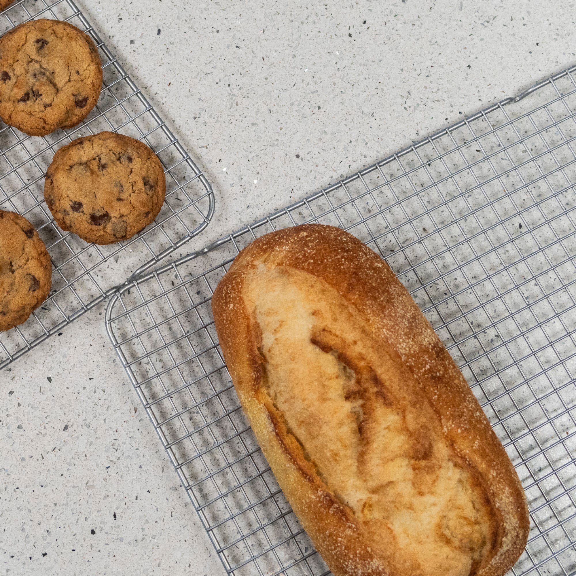 A loaf of bread and chocolate chip cookies cool on the Viking 2-pc Cooling Rack Set by Viking Culinary Products, which fits a Viking 17-inch baking sheet, atop a light-colored countertop.
