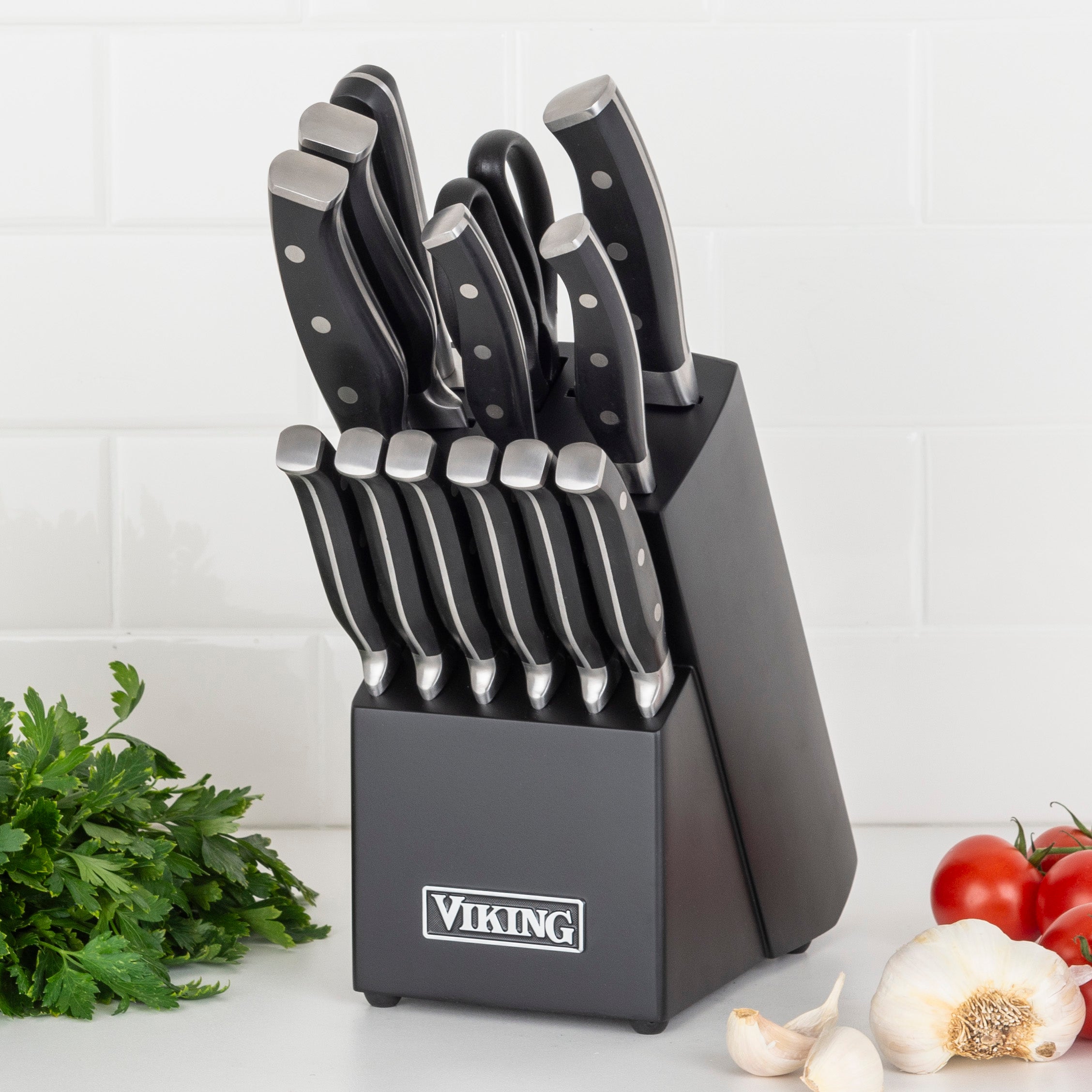 The Viking 14-Piece X50CrMoV15 Steel Cutlery Set with Black Block by Viking Culinary Products sits on a white countertop, surrounded by fresh parsley, garlic cloves, and tomatoes.