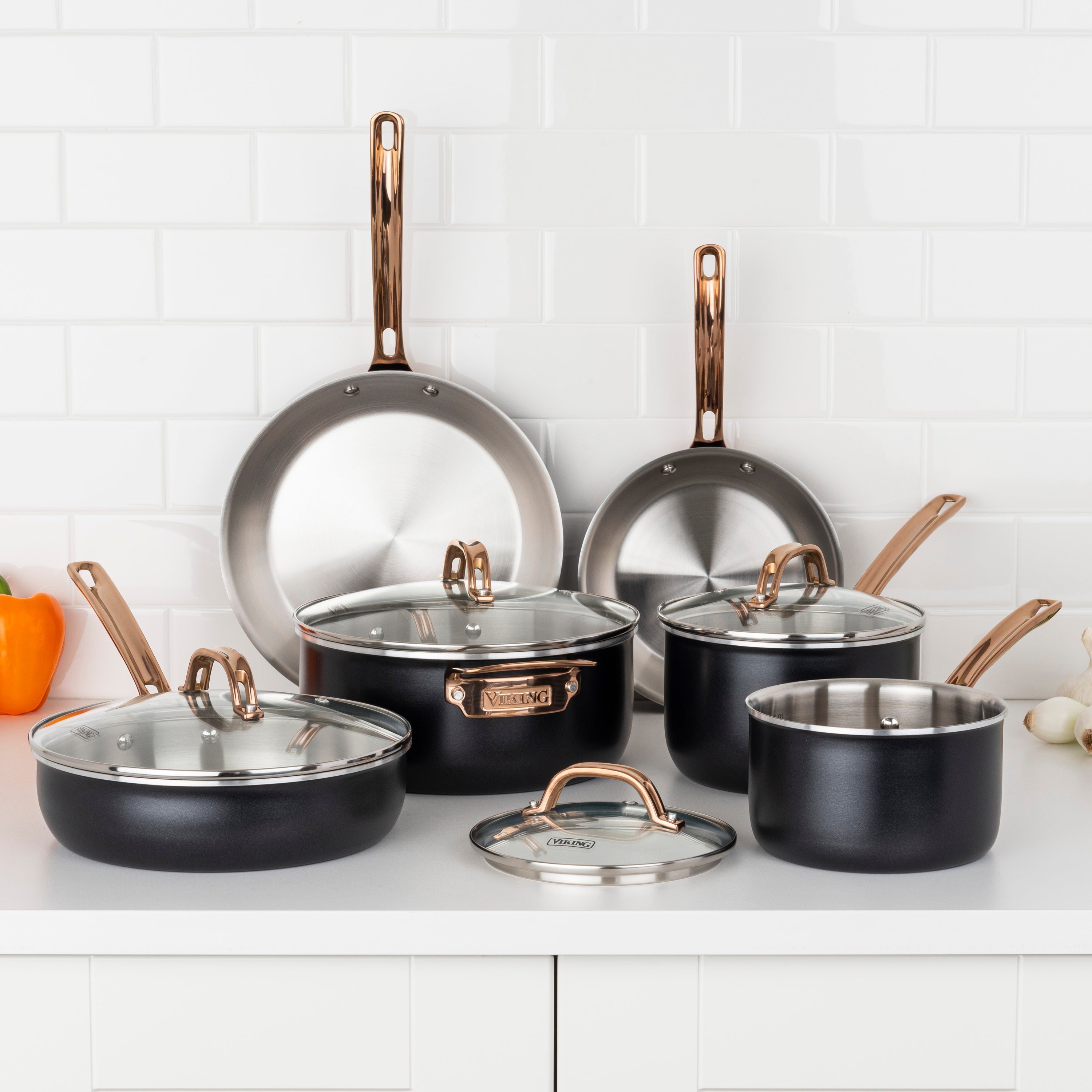 The Viking 10pc 3-Ply Black & Copper Stainless Steel Cookware Set by Viking Culinary Products is displayed on a white kitchen counter with a subway tile backsplash; some pieces have glass lids, and vegetables sit nearby.