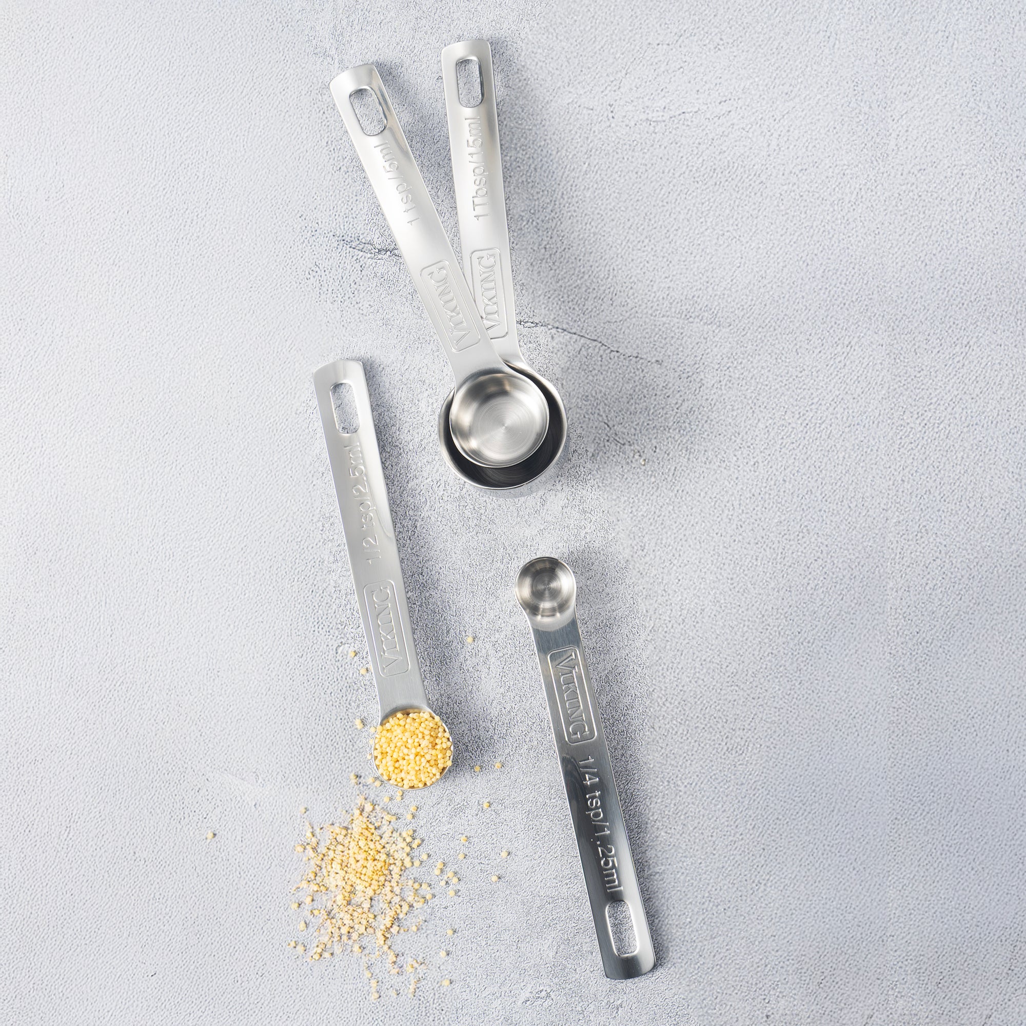 The Viking 4pc Stainless Steel Measuring Spoon Set by Viking Culinary Products is arranged on a light gray surface, with one spoon holding millet and some spilled nearby. The nesting spoons feature labeled measurements and are dishwasher safe.