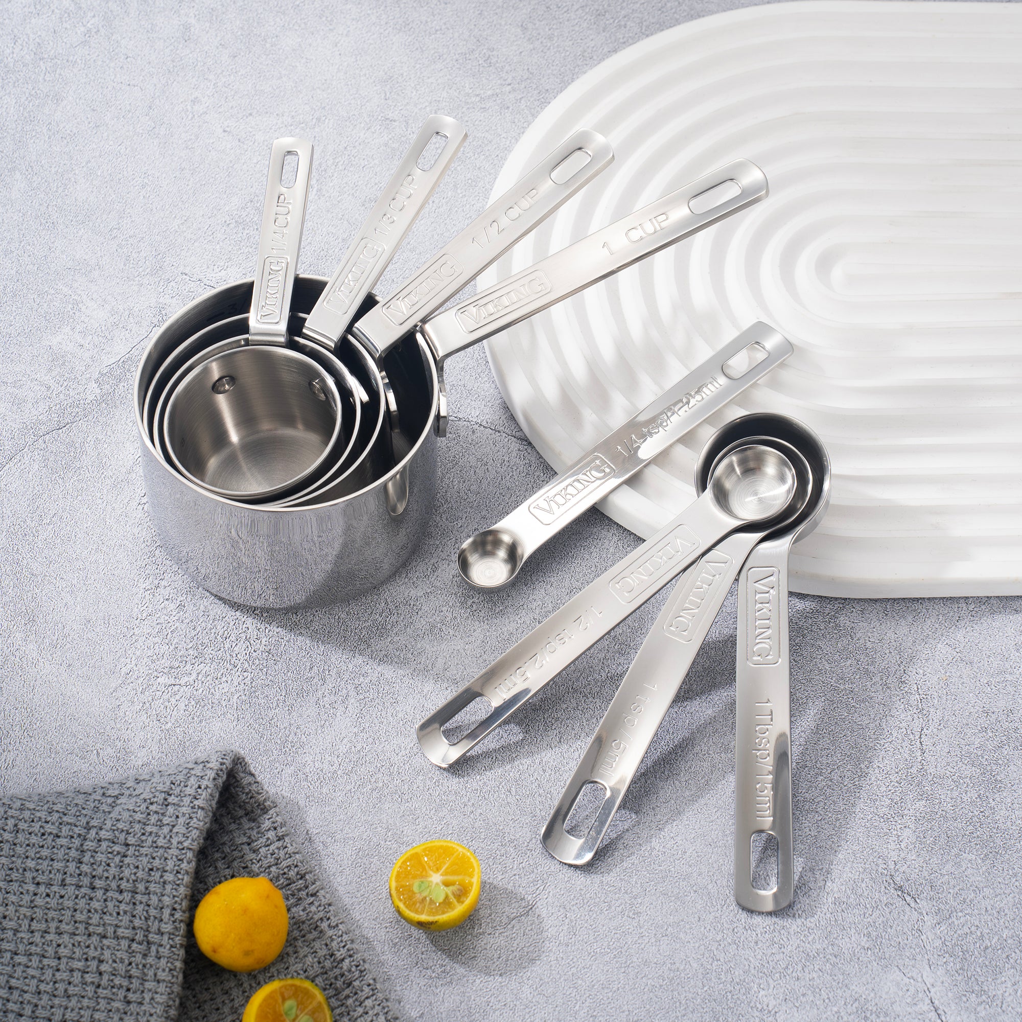 The Viking Stainless Steel Measuring Cup Set by Viking Culinary Products is arranged on a gray surface with a white tray, gray cloth, and halved lemons. These durable measuring cups are dishwasher safe for easy cleaning.