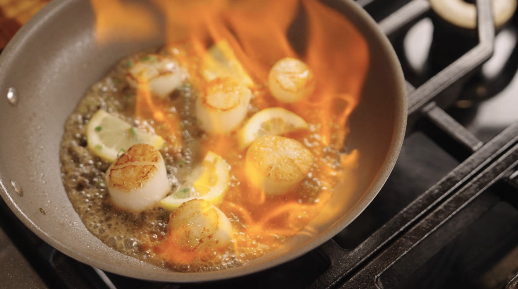 Scallops and lemon slices are being flambéed in a skillet on a stovetop, with visible flames and bubbling butter surrounding the food.