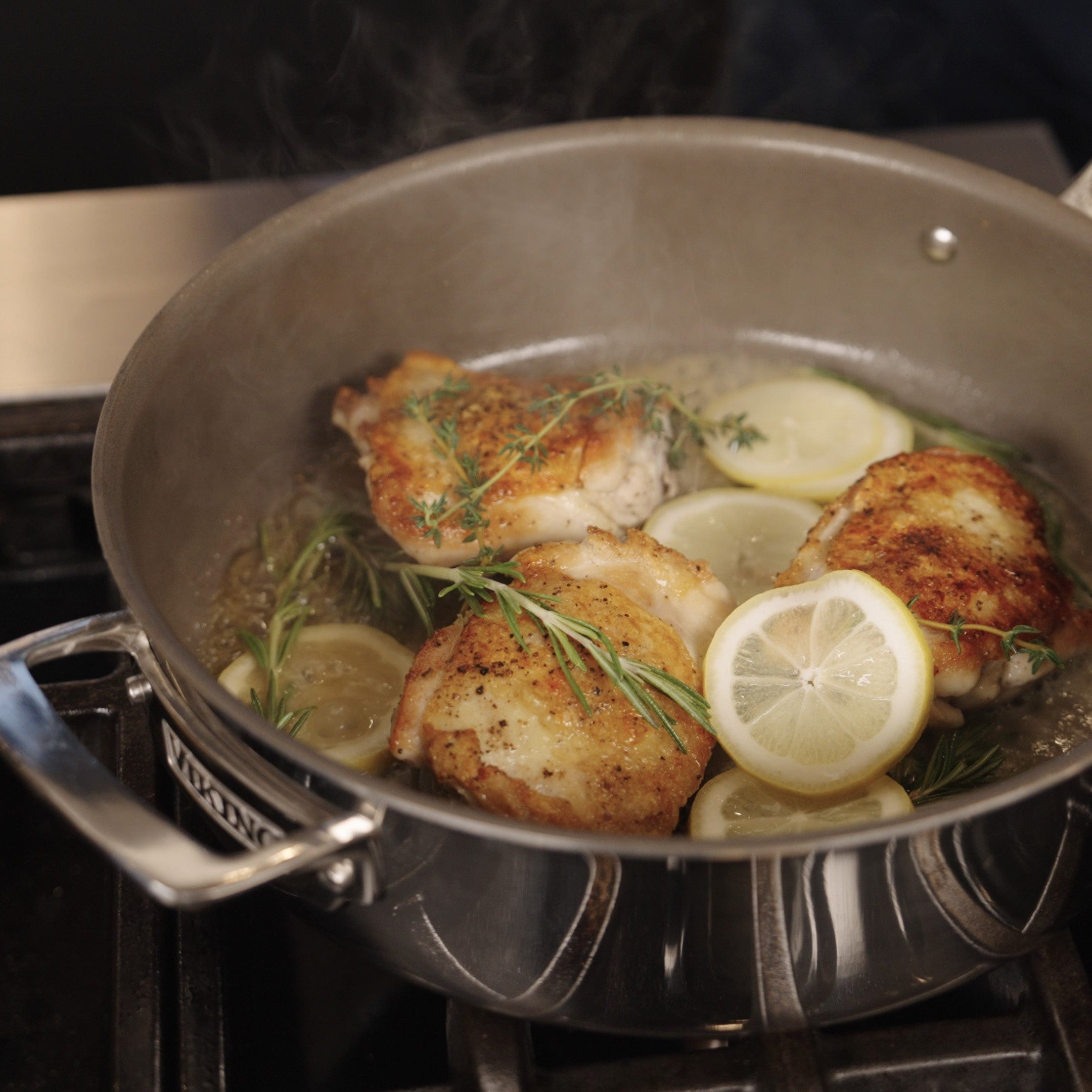 Chicken thighs sizzle in the Viking PureGlide Pro 3-Ply 4.5-Quart Saute Pan with rosemary, thyme, and lemon slices from Viking Culinary Products, sending up steam as they cook under the metal lid.