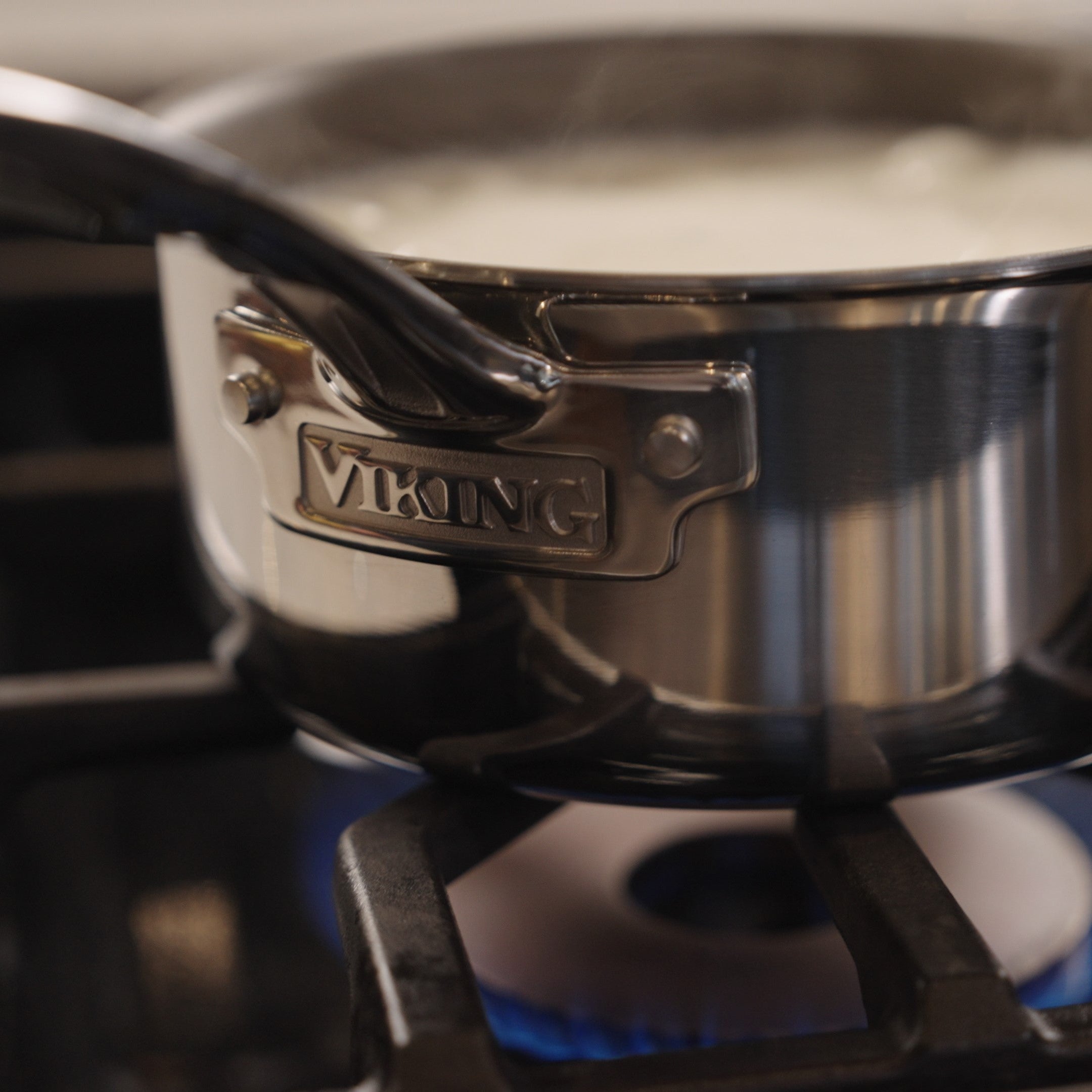 A Viking PureGlide Pro 3-Ply 2-Quart Sauce Pan with Metal Lid by Viking Culinary Products is heating on a gas stove, blue flames visible underneath and liquid simmering inside.
