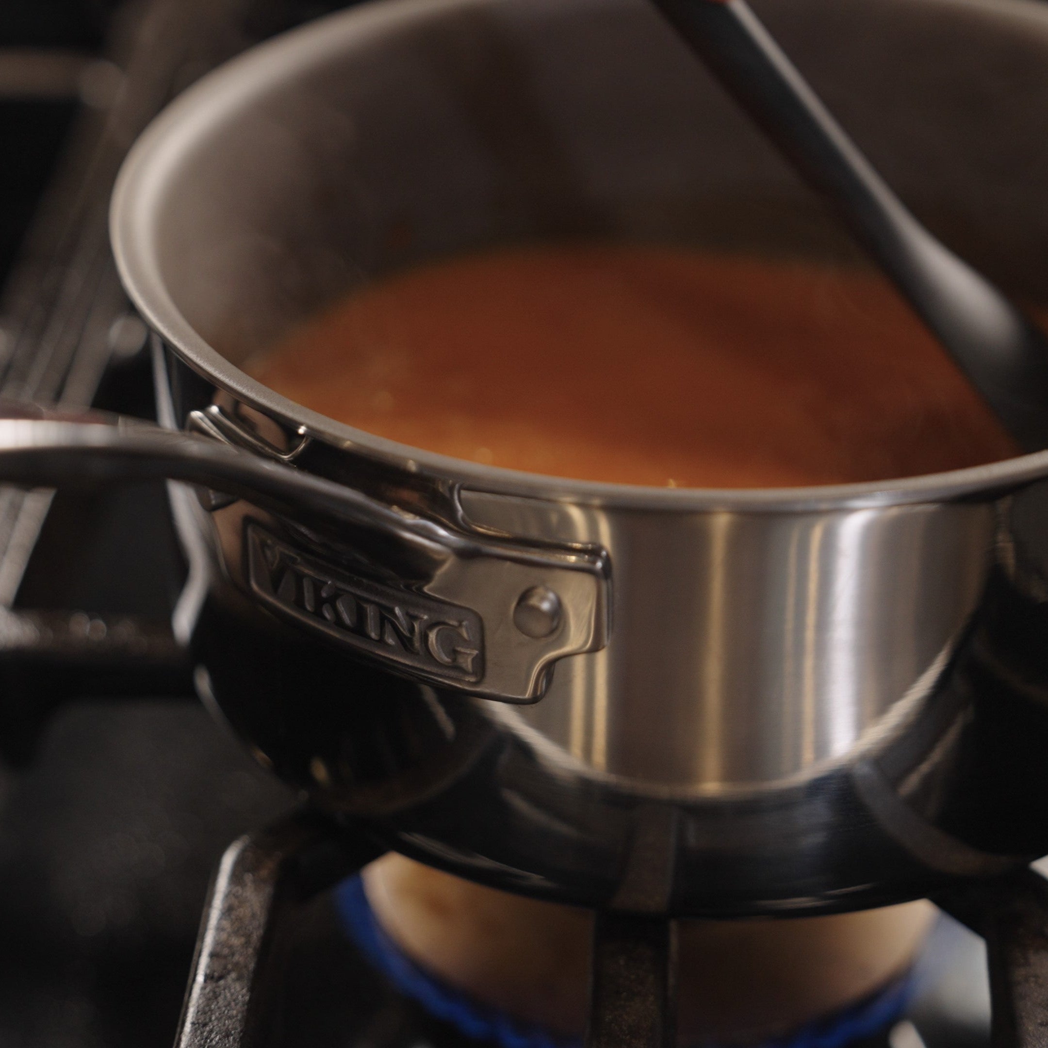 A Viking Culinary Products PureGlide Pro 3-Ply 3-Quart Sauce Pan with metal lid simmers a reddish-brown liquid on a blue-flamed burner, steam rising as a black spoon stirs the contents.