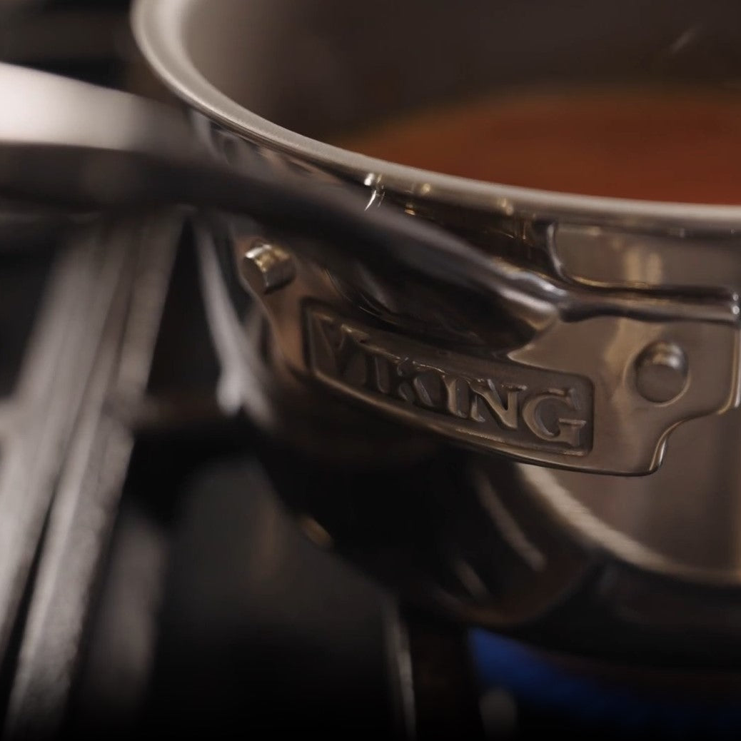 Close-up of a Viking PureGlide Pro 3-Ply 2-Quart Sauce Pan with Metal Lid by Viking Culinary Products on a stovetop, filled with reddish liquid. The VIKING logo is engraved on the handle, with blue flames visible beneath.
