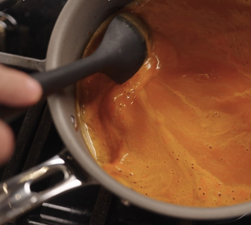 A hand stirs a thick, creamy orange sauce in a Viking PureGlide Pro 3-Ply 3-Quart Sauce Pan with Metal Lid from Viking Culinary Products using a black spoon on the stovetop.