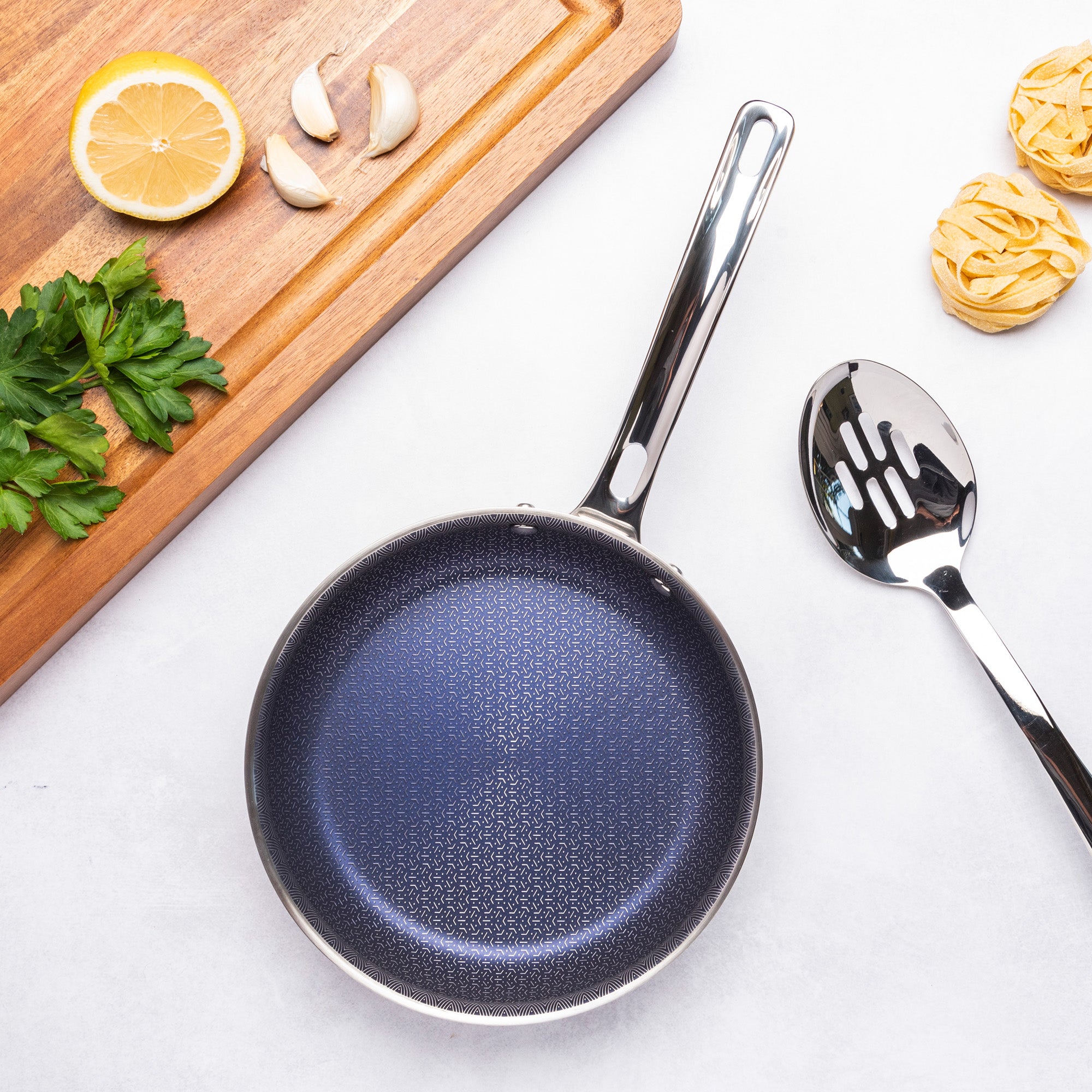 The Viking 3-Ply Hybrid Plus 8-inch Nonstick Fry Pan by Viking Culinary Products, featuring an induction-ready patterned interior, rests on a white surface beside a slotted spoon and a board with lemon, garlic, parsley, and pasta nests.