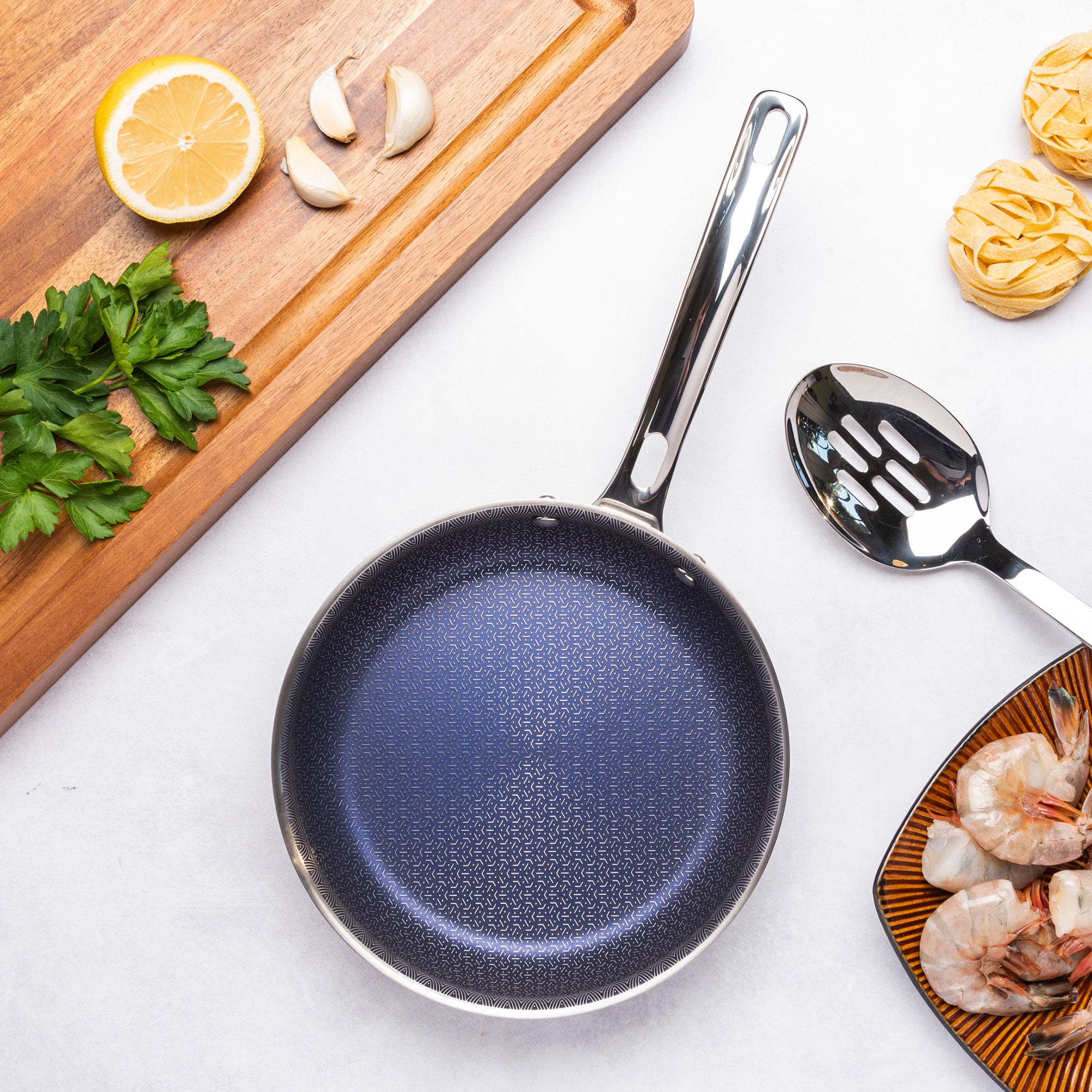 The Viking Culinary Products Viking 3-Ply Hybrid Plus 8-inch Nonstick Fry Pan with a fast-heating, induction-ready base rests on a white surface, surrounded by a slotted spoon, raw shrimp, pasta nests, parsley, garlic cloves, and lemon on a wooden board.