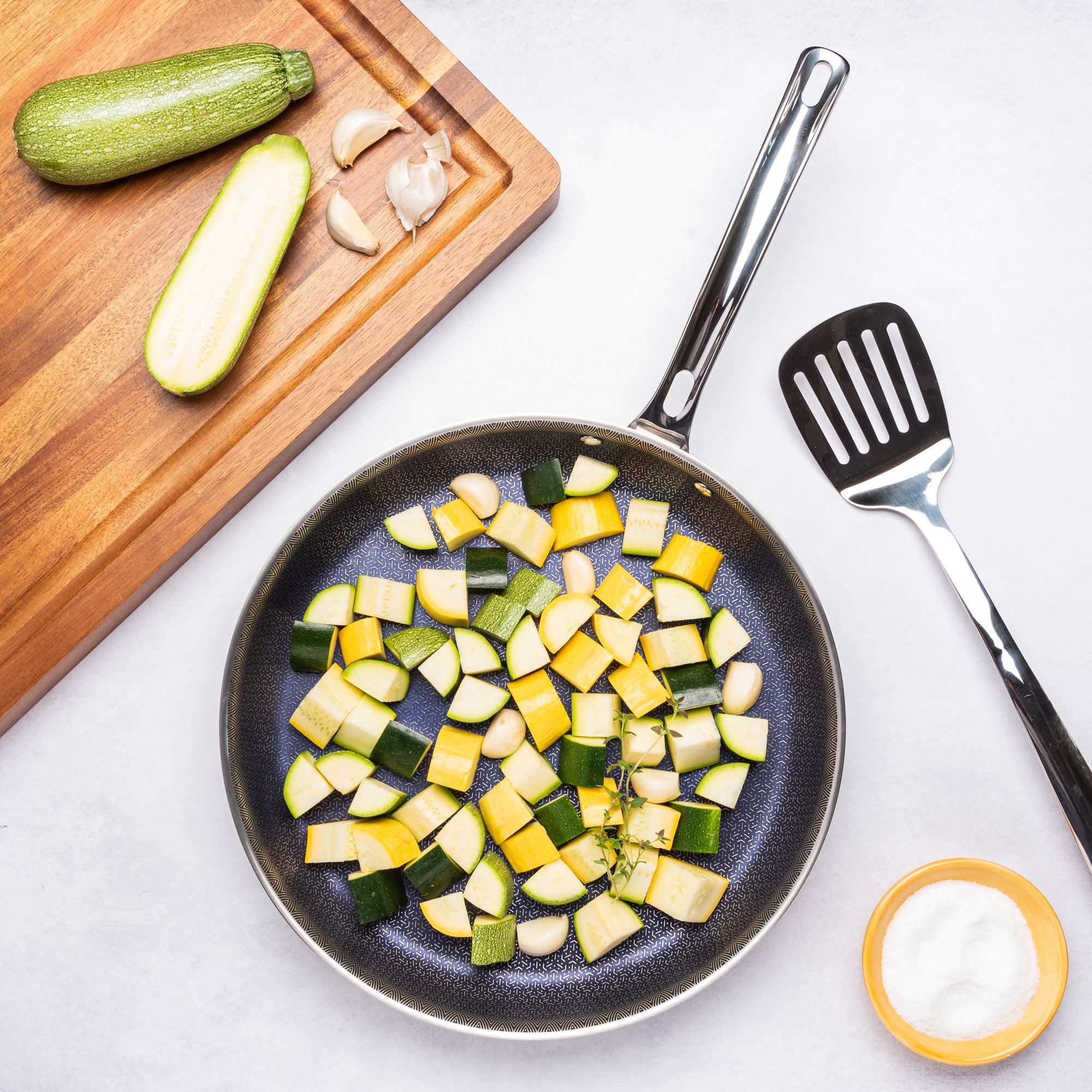 The Viking Culinary Products 3-Ply Hybrid Plus 12-inch Nonstick Fry Pan cooks diced yellow and green zucchini next to a spatula, salt bowl, cutting board with whole and halved zucchini, and garlic on a white surface.