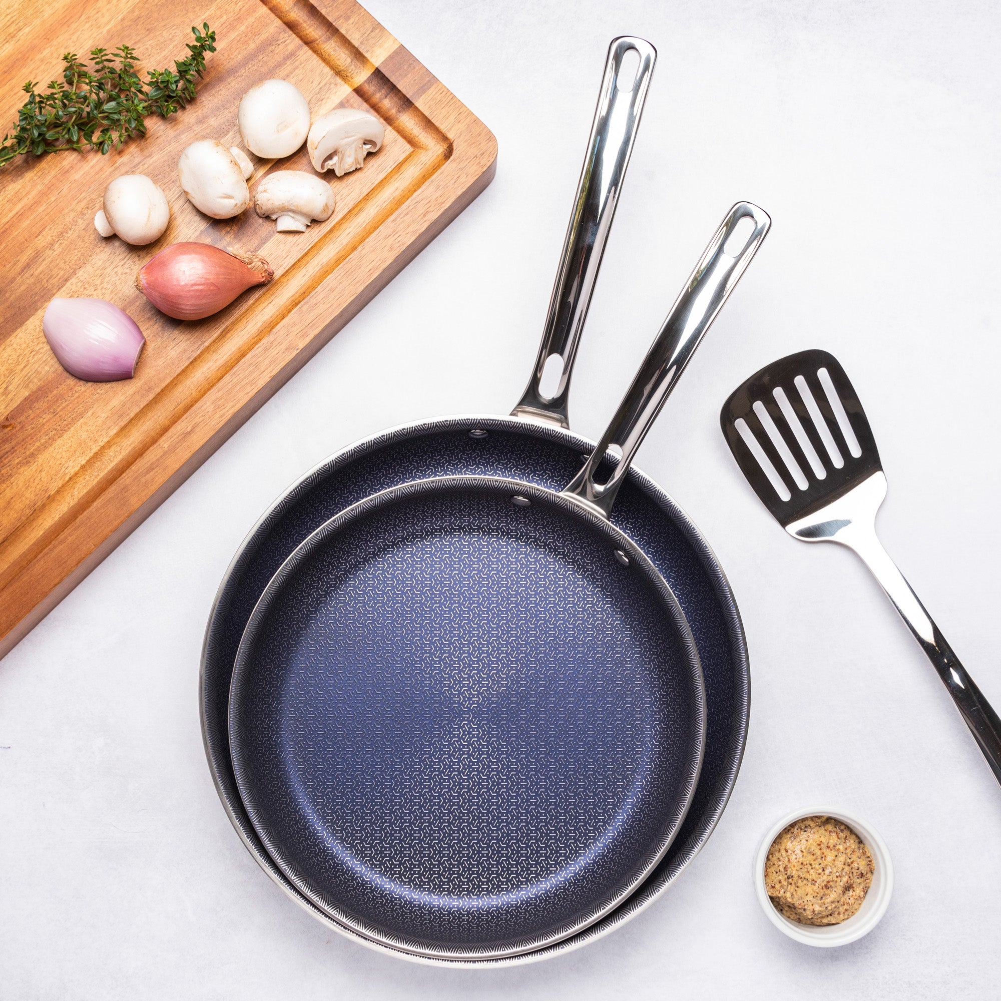 The Viking 3-Ply Hybrid Plus 2 Pc Nonstick Fry Pan Set (9.5 & 11) from Viking Culinary Products is stacked on a white surface with a spatula, seasoning bowl, and a wooden board holding mushrooms, shallots, and fresh thyme.