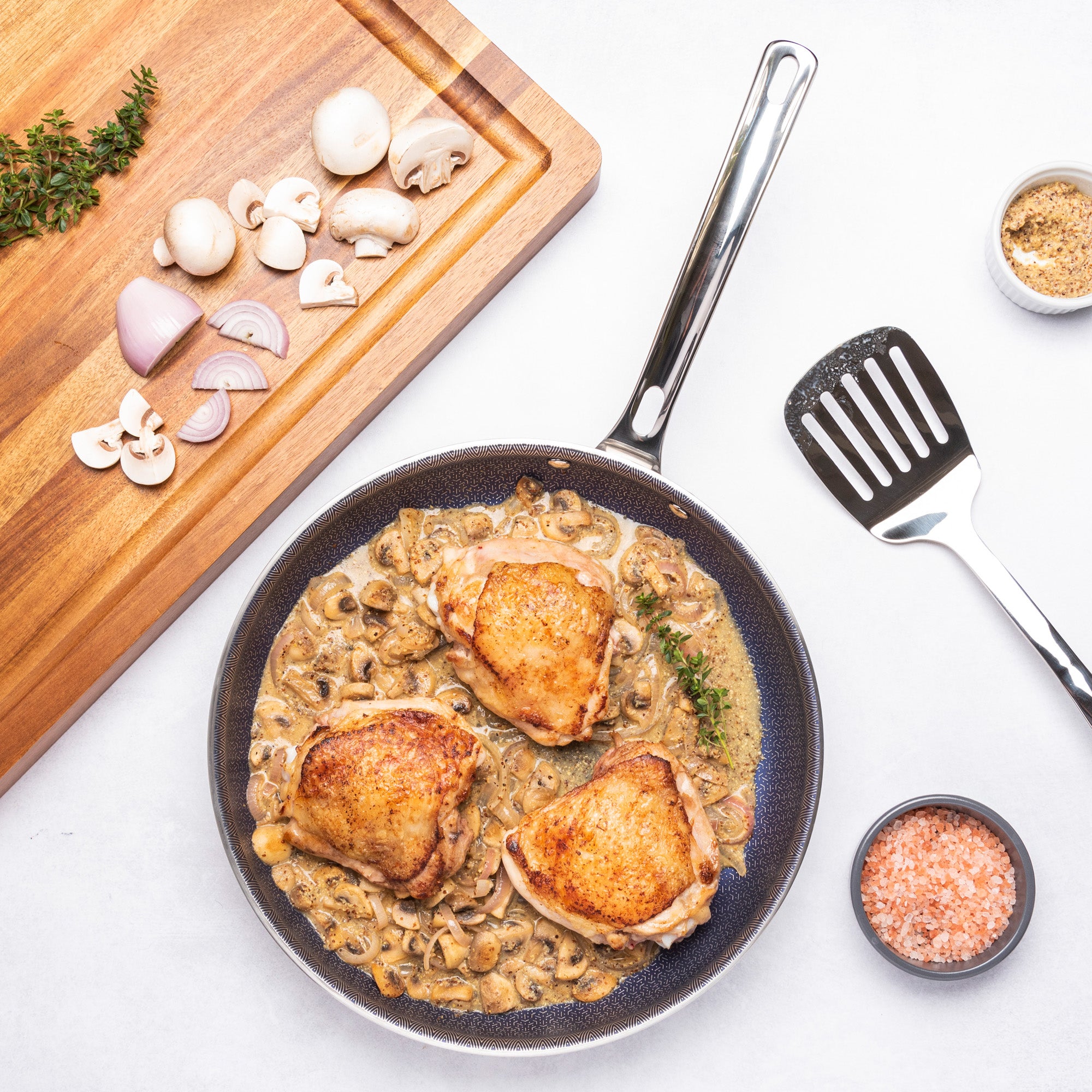 The Viking 3-Ply Hybrid Plus 2 Pc Nonstick Fry Pan Set by Viking Culinary Products cooks creamy mushroom chicken thighs and sits beside an ergonomic spatula, pink salt, seasonings, and a board with mushrooms, shallots, and herbs.