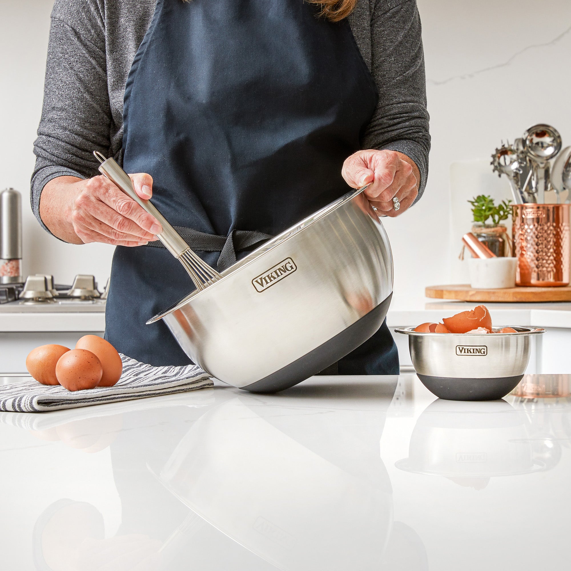 A person in a dark apron uses a Viking 10-Piece Stainless Steel Mixing Bowl with a silicone base on the counter, surrounded by eggs and another bowl. Cooking utensils and a copper container are visible in the background.