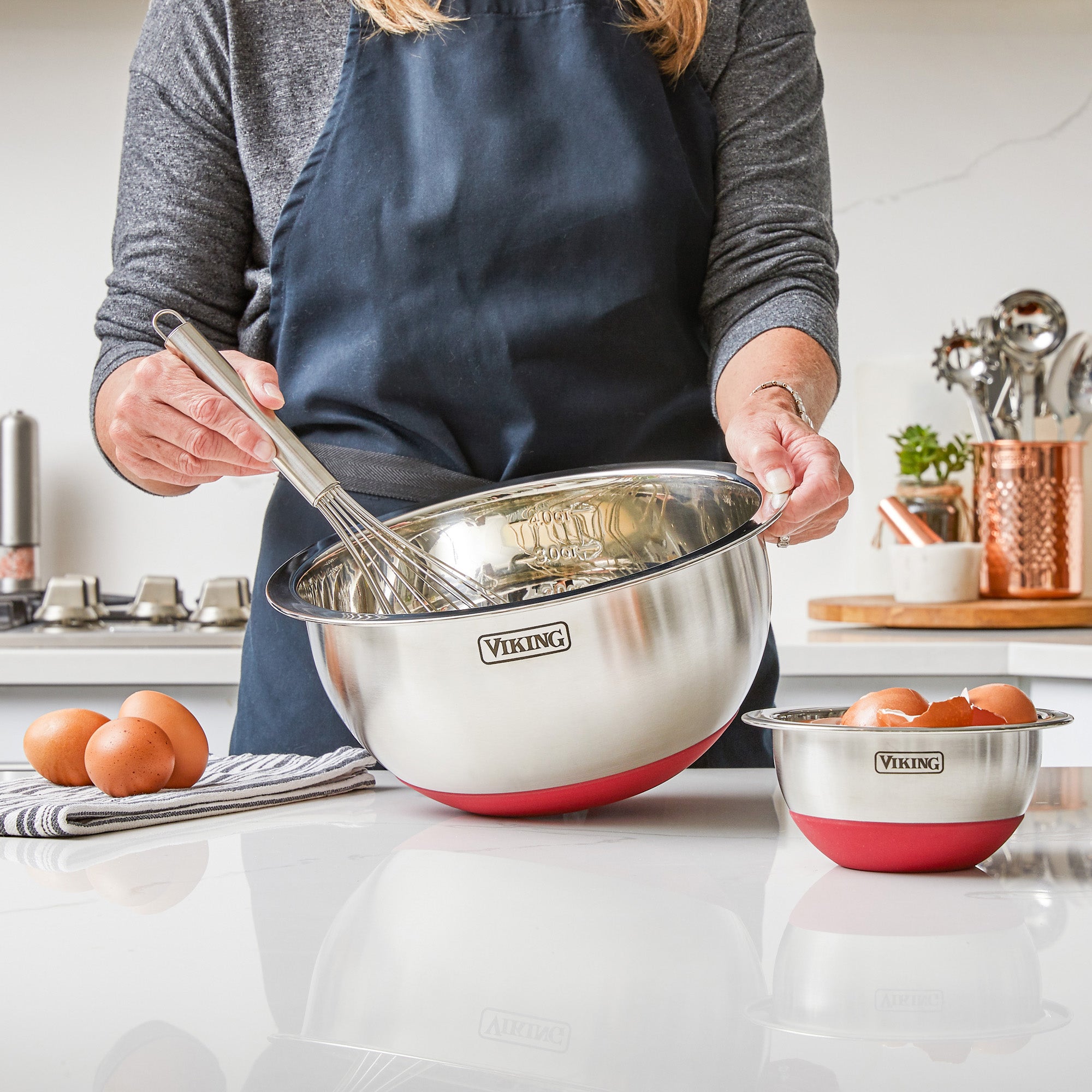 A person in a navy apron holds a whisk and a large red Viking Culinary Products 10-Piece Stainless Steel Mixing Bowl with silicone base, while eggs rest on a striped towel beside a smaller bowl from the set on the kitchen counter.