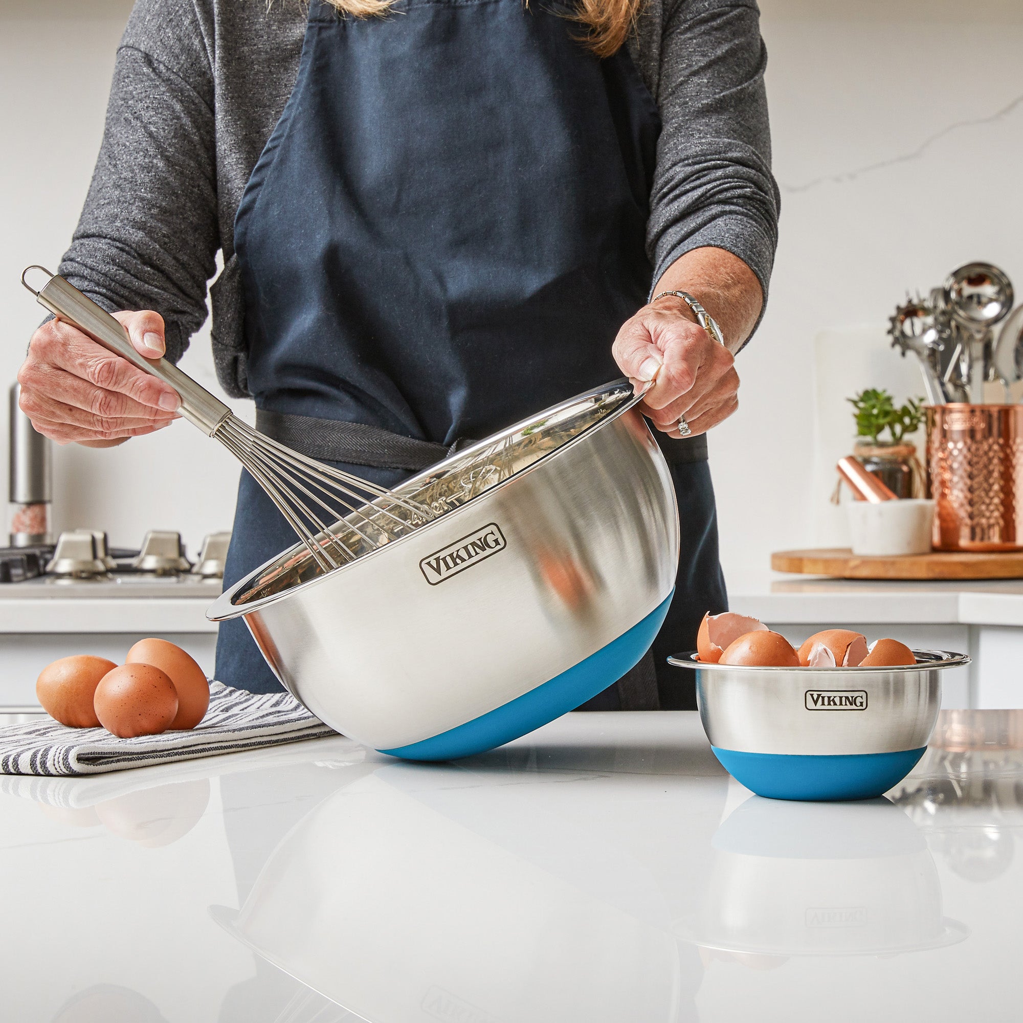 Wearing a dark apron, a person whisks ingredients in a Viking 10-Piece Stainless Steel Mixing Bowl with a teal silicone base from Viking Culinary Products, with eggs and another bowl from the set on the kitchen counter.