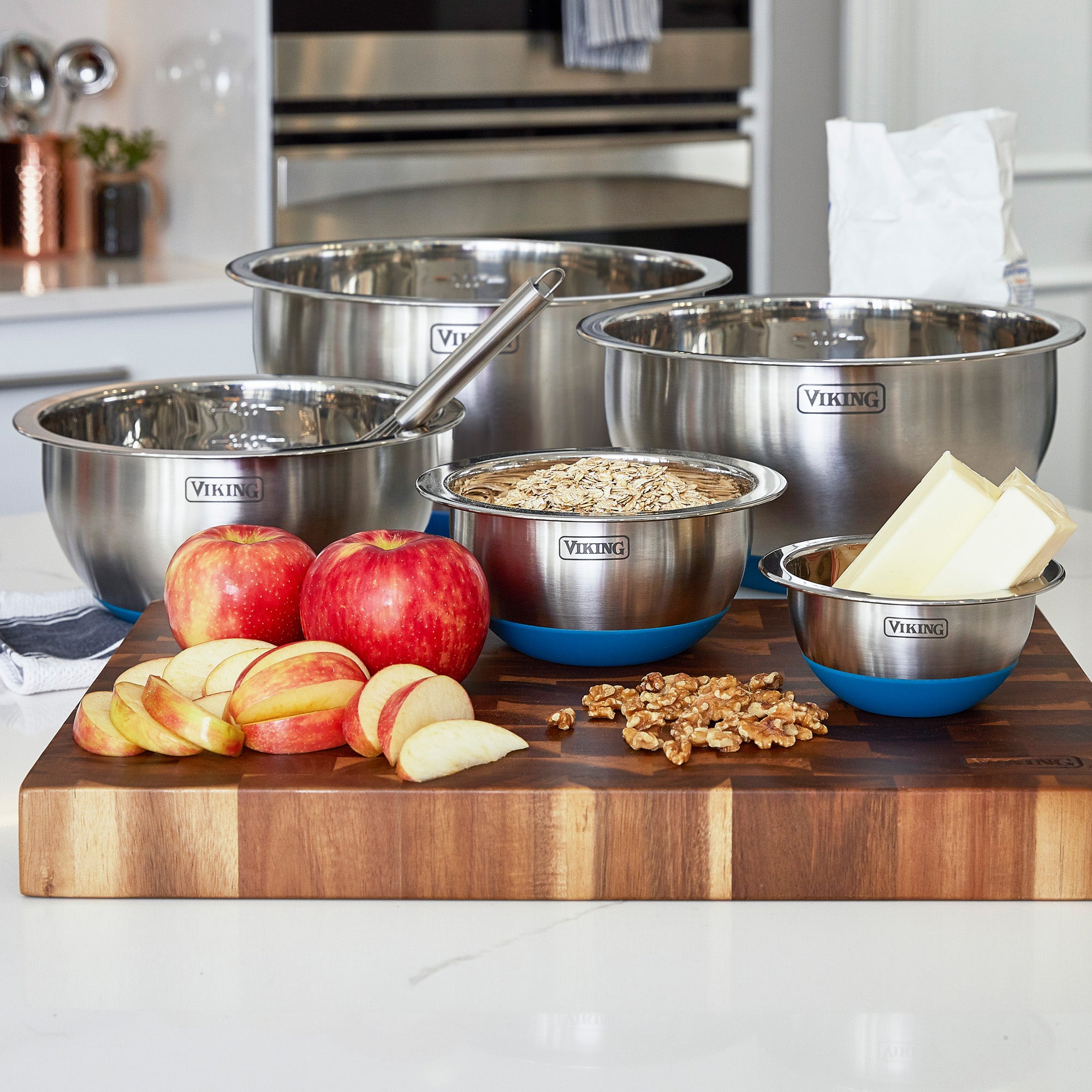 A Viking Culinary Products 10-Piece Stainless Steel Mixing Bowl Set with teal lids sits on the counter, with apples, apple slices, walnuts, butter sticks, and a bowl of oats arranged on a wooden cutting board in front.