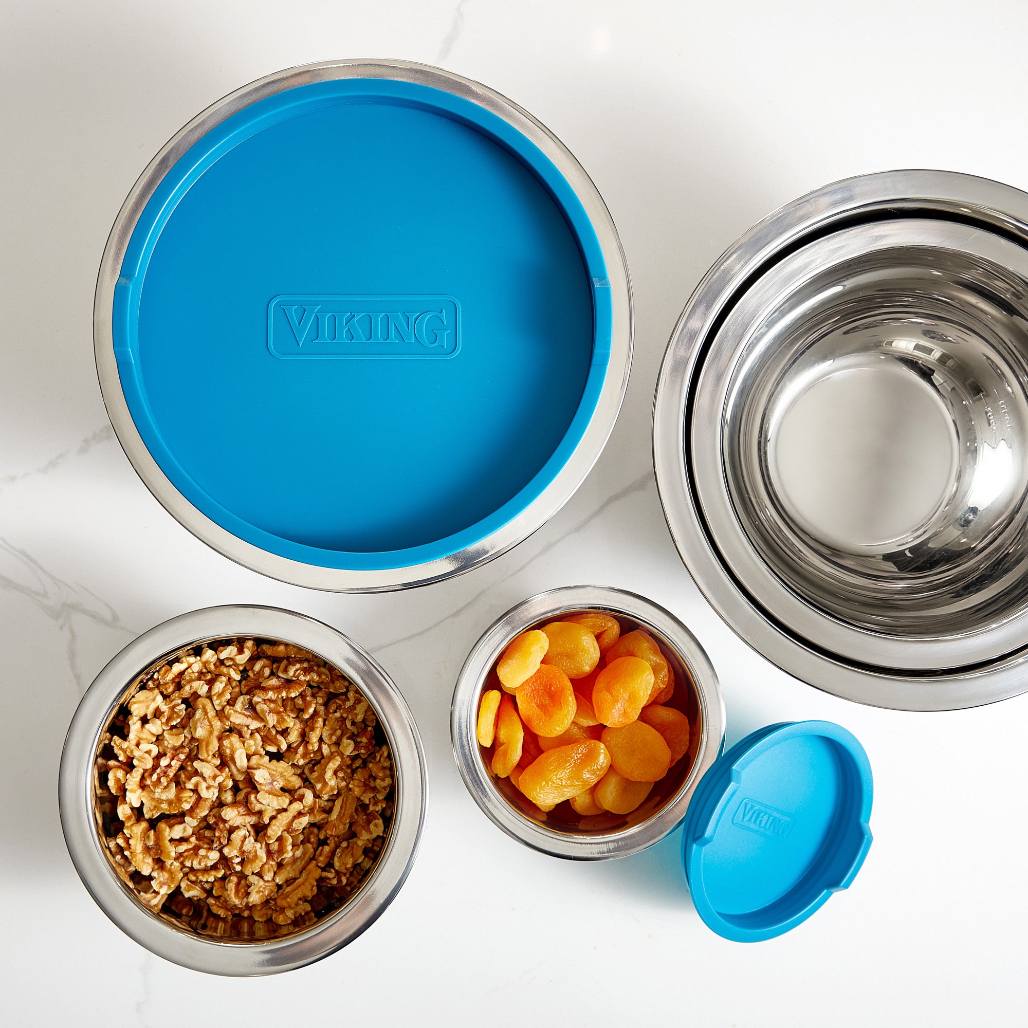 Four Viking Culinary Products stainless steel mixing bowls from the 10-piece set with teal lids are displayed on a white surface; one holds walnuts, another dried apricots, while two are empty and ready for use.