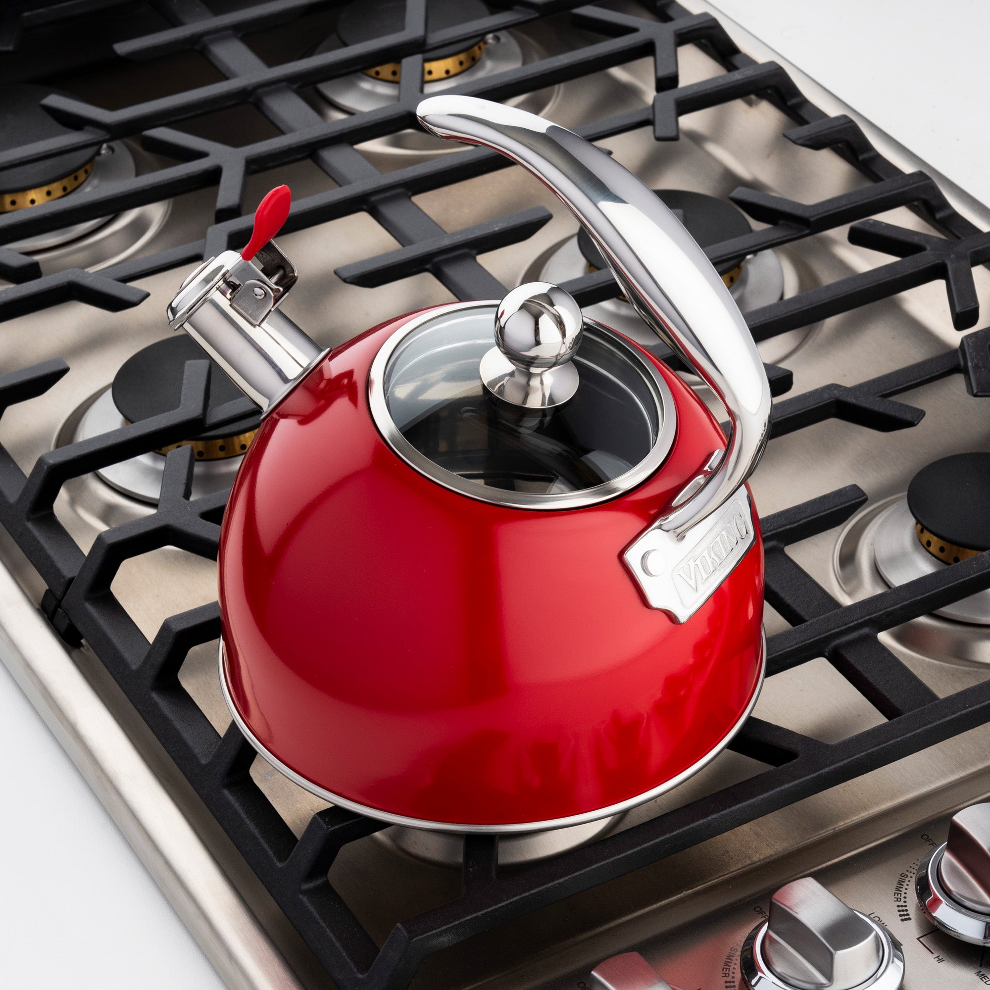 A Viking 2.6-Quart Red Stainless Steel Whistling Kettle by Viking Culinary Products with a silver handle and lid sits on a stainless steel gas stove with black grates. The spout lever is up, ready to whistle, and the stove has multiple burners.